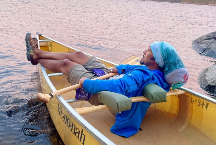 Derick Lugo rests in a canoe on a lake wearing Oboz trail sandals.  