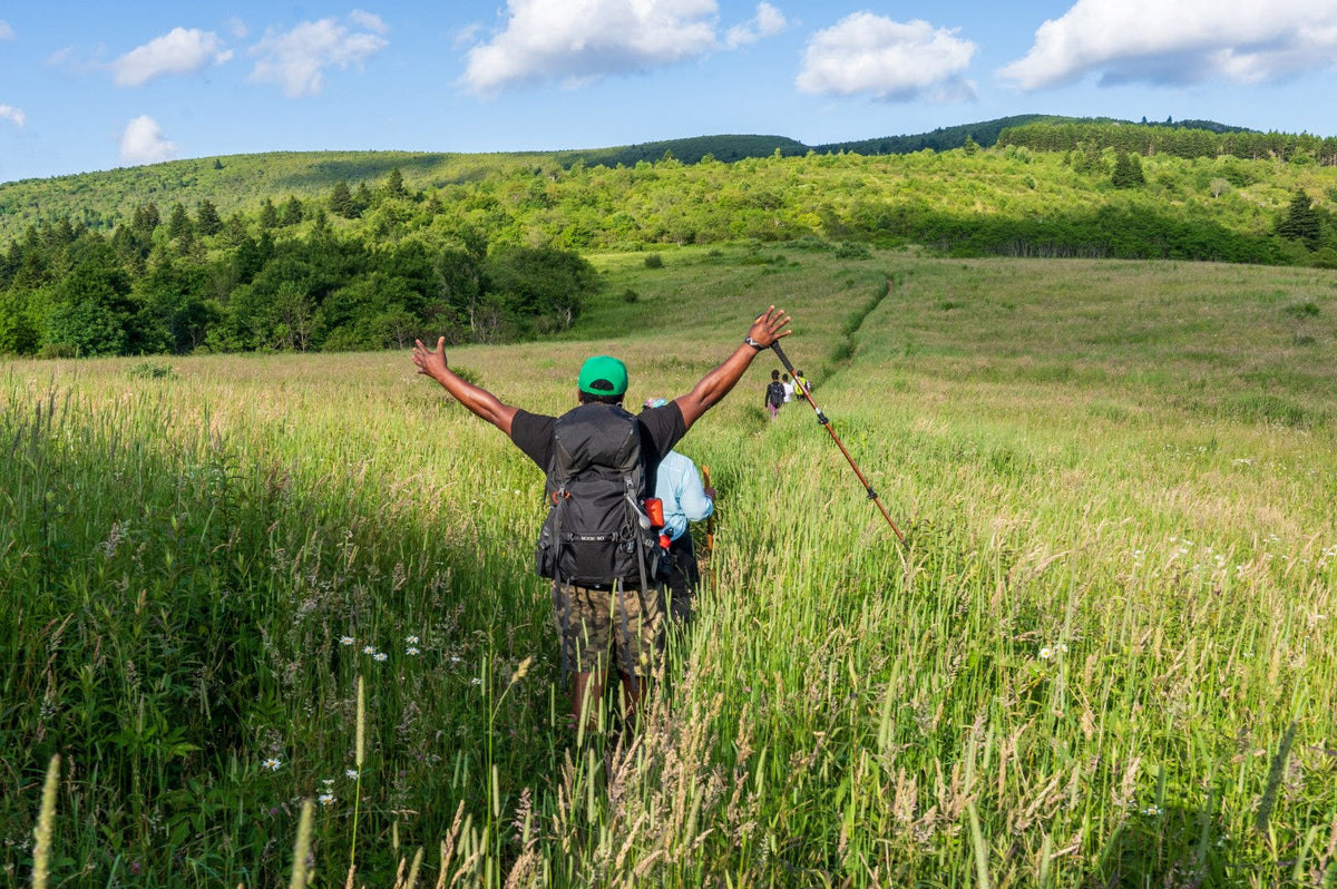 Hiker is raising hands victoriously while walking through a grassy plain.