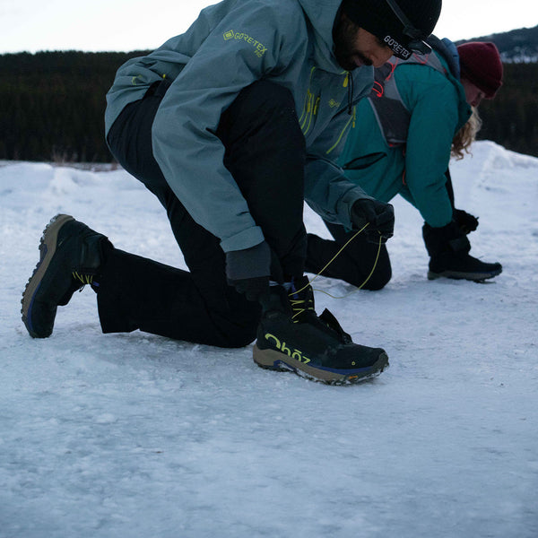 A close-up of a person's legs stepping into a muddy puddle outdoors, wearing black Oboz Katabatic Storm winter boots with yellow and blue accents, rainbow-striped socks, and black leggings with a white stripe.