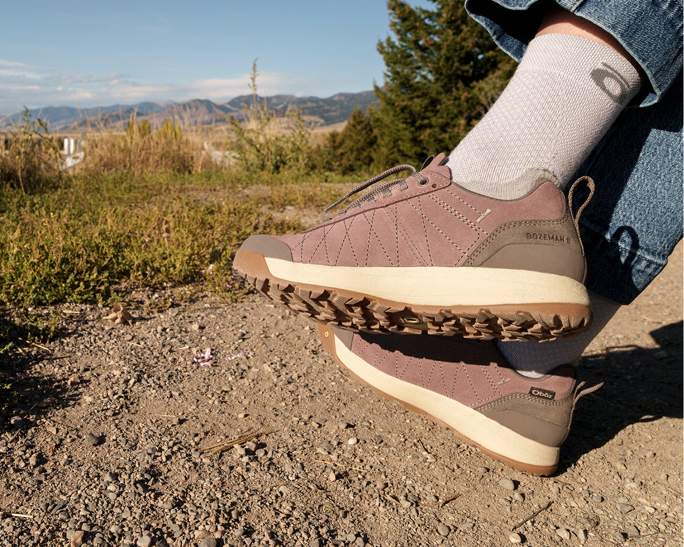 A person outdoors wearing a pair of women's Bozeman II shoes on a gravel path with mountains and trees in the background.