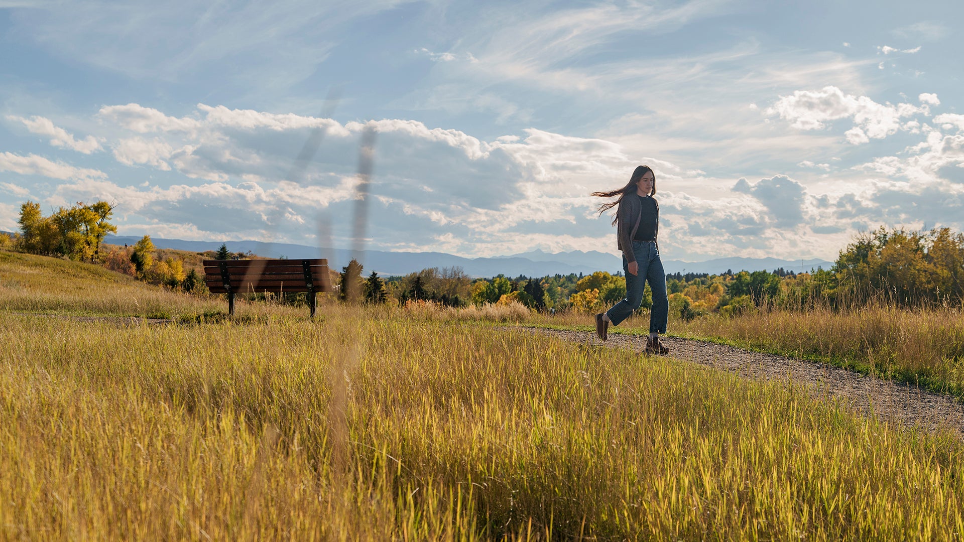 A woman walks along a gravel path in a grassy field under a cloudy blue sky, with trees and a bench in the background.