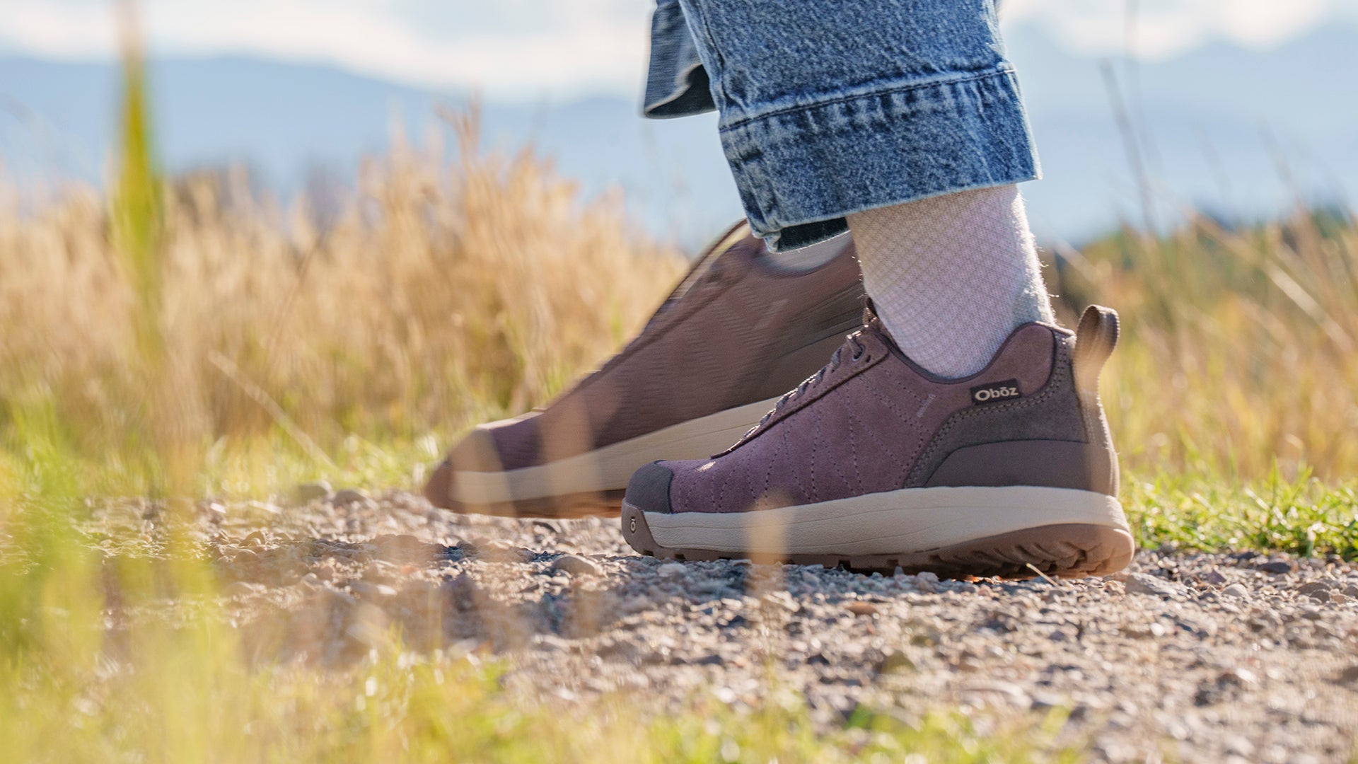 A person wearing Fig Oboz Bozeman II shoes with white socks, standing on a gravel path in a grassy outdoor setting.