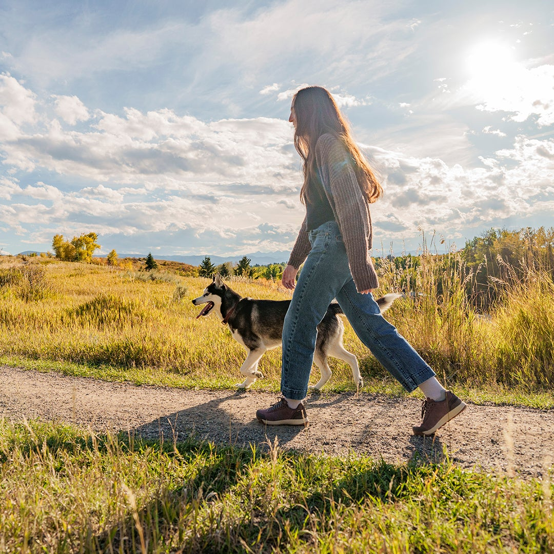 A woman walks on a dirt path with a husky dog in an open grassy field under a bright sky with clouds.