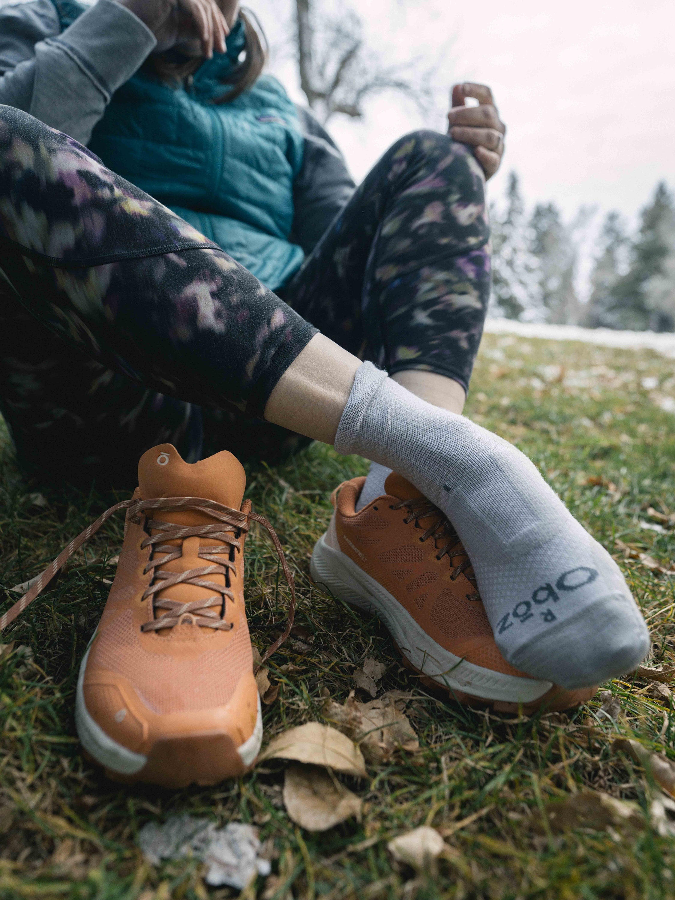 Person sitting on grass wearing orange Oboz Katabatic LT hiking shoes, camouflage pants, and white Oboz socks.