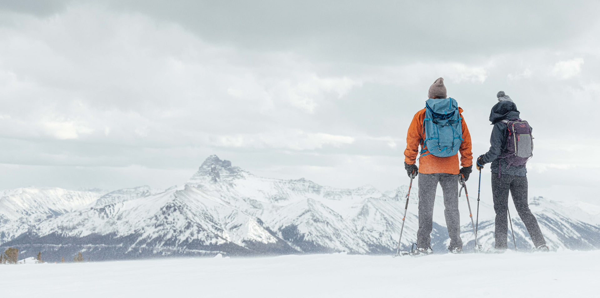 Two people with snowshoes and backpacks standing on a snowy landscape with mountains in the background