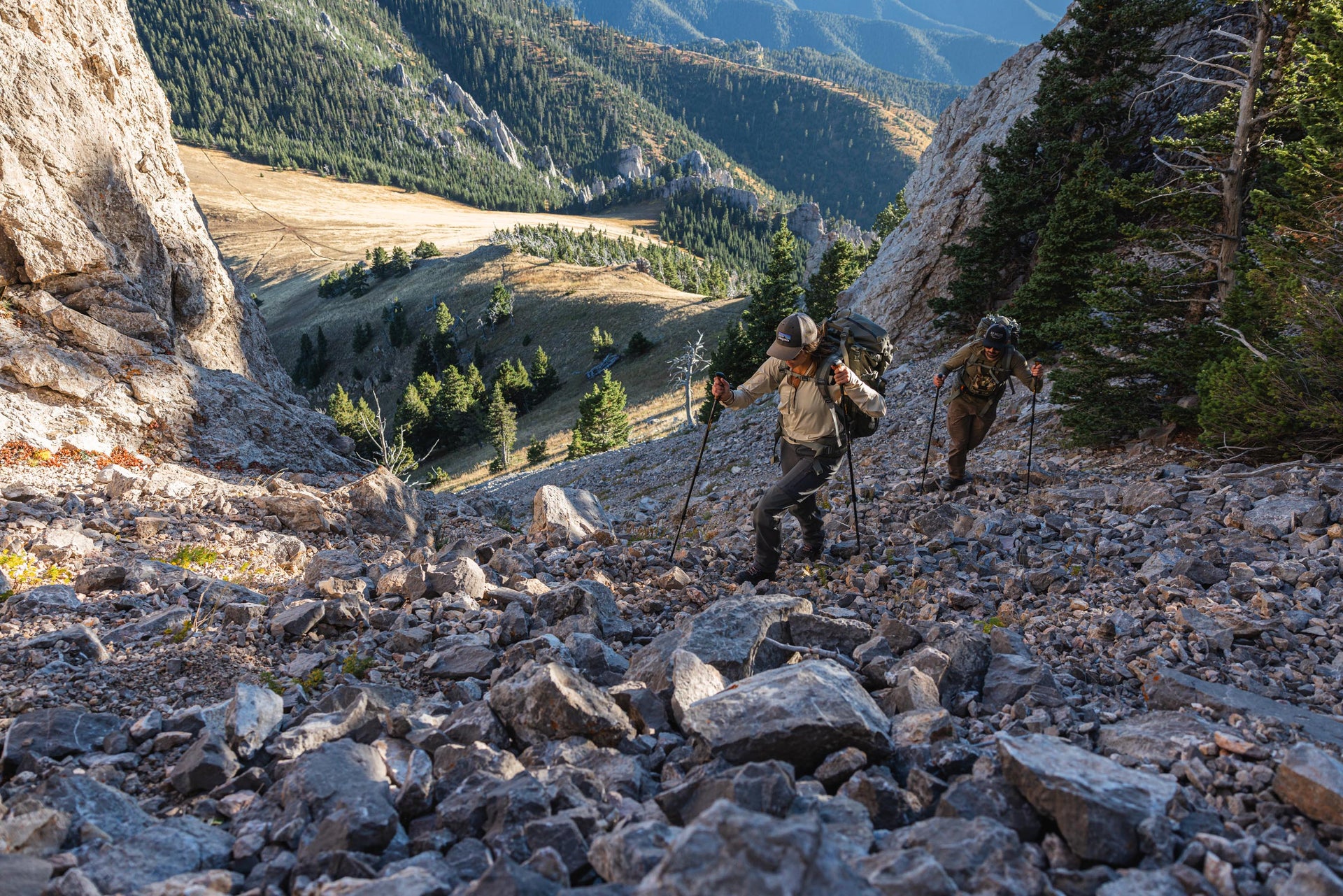 Two hikers on a rocky mountain trail with a scenic mountain landscape.