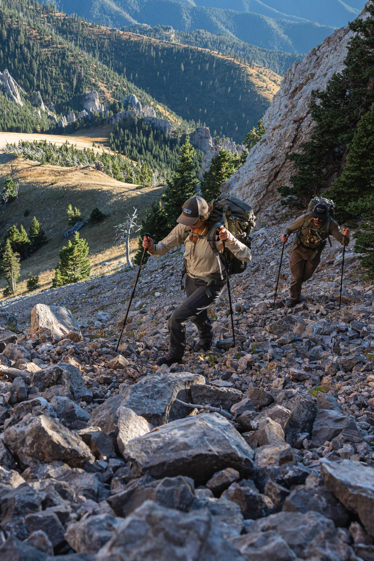 Two hikers on a rocky mountain trail with a scenic mountain landscape.