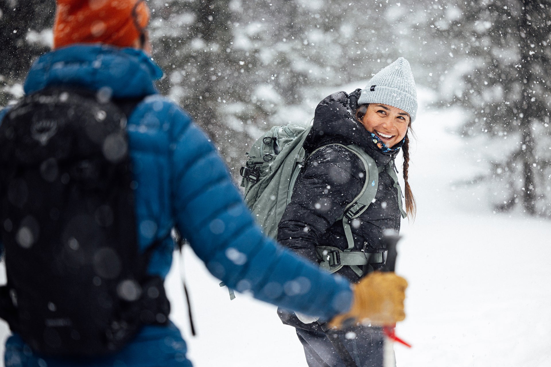 Two people in winter clothing smiling in a snowy forest during a snowstorm.