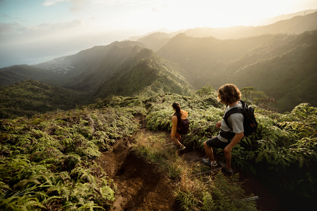 Two hikers sitting on a mountain trail with a scenic view of mountains and greenery.