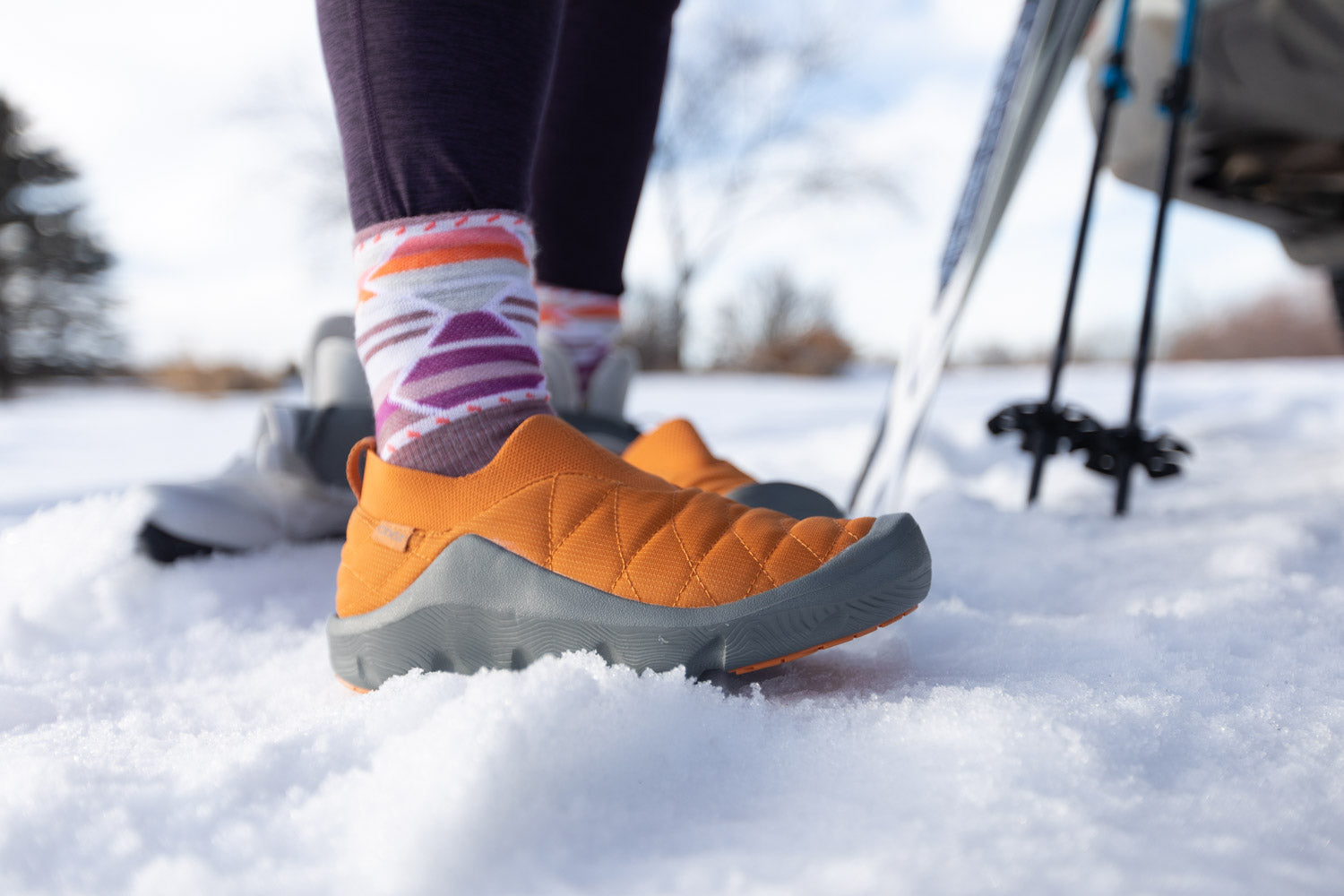 Person wearing orange Oboz Whakata Puffy slippers and colorful socks standing in the snow with skis in the background.