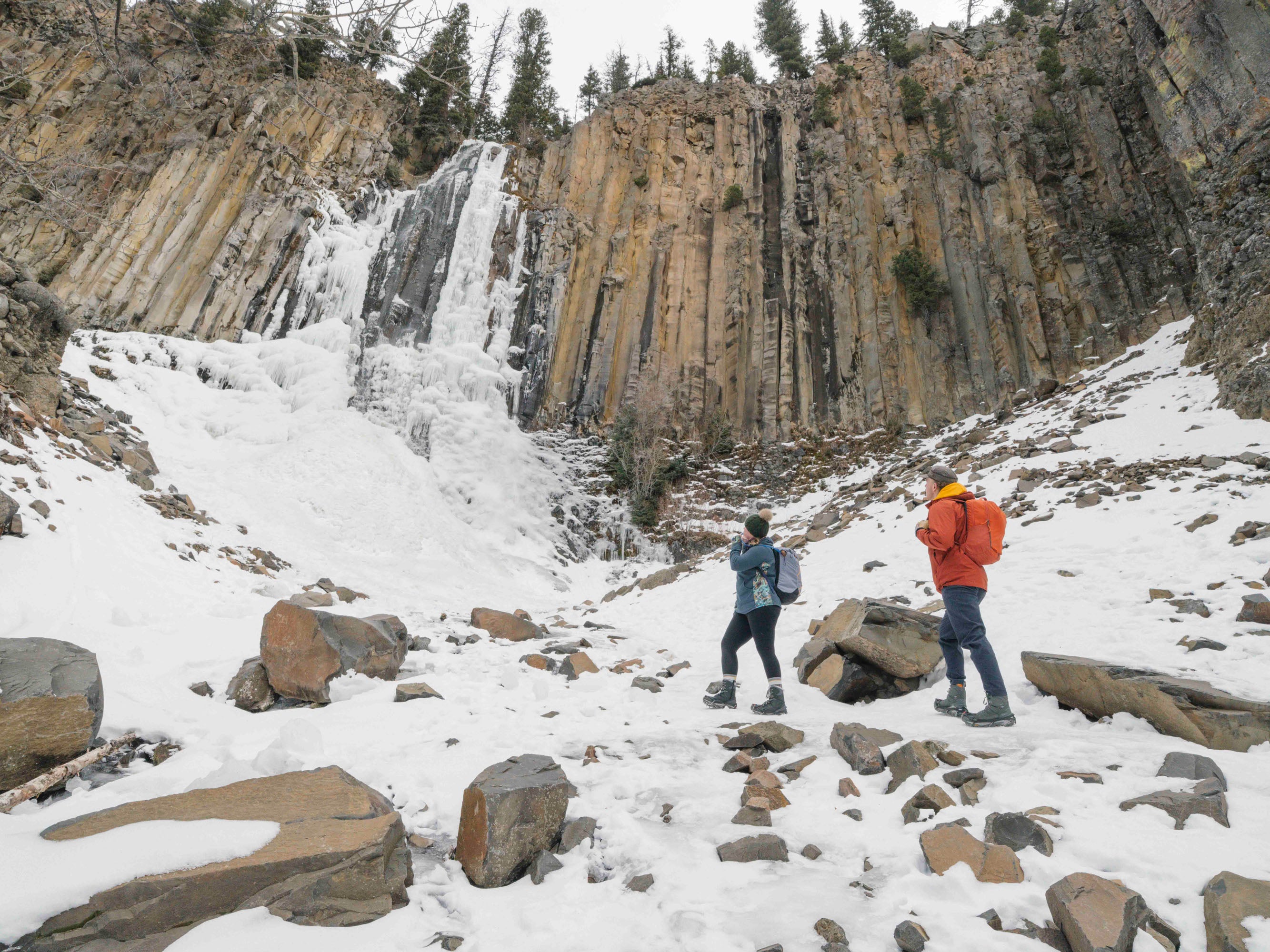 Two people hiking near a frozen waterfall in a snowy landscape.