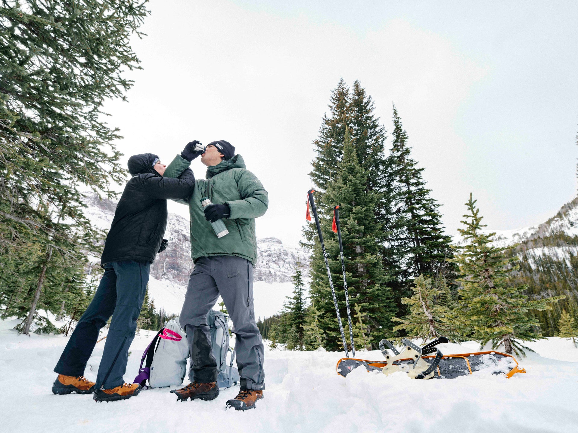 Two people in winter gear celebrating with tea in a snowy forest setting.