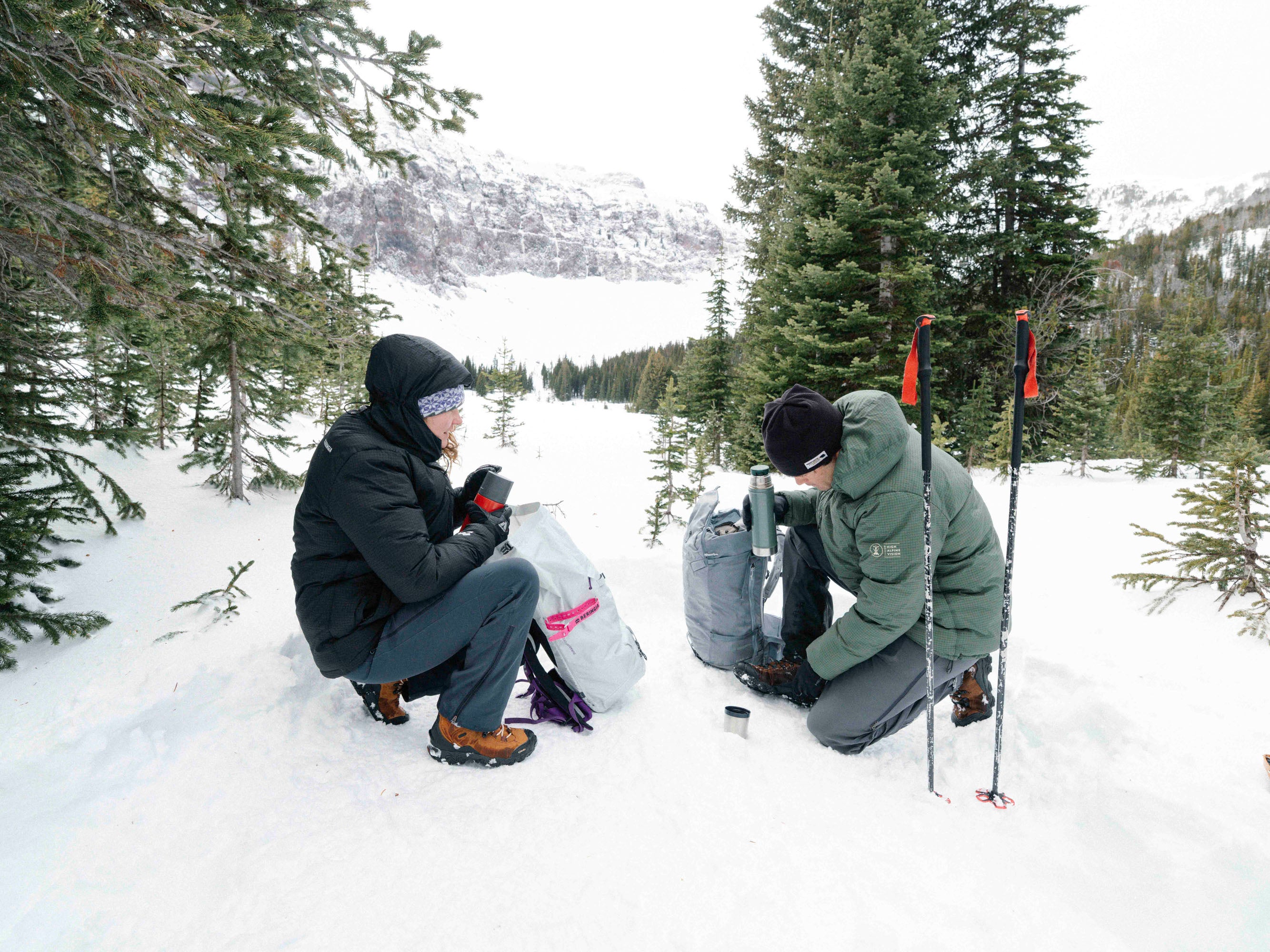 Two people in winter gear taking on break while hiking in a snowy forest.