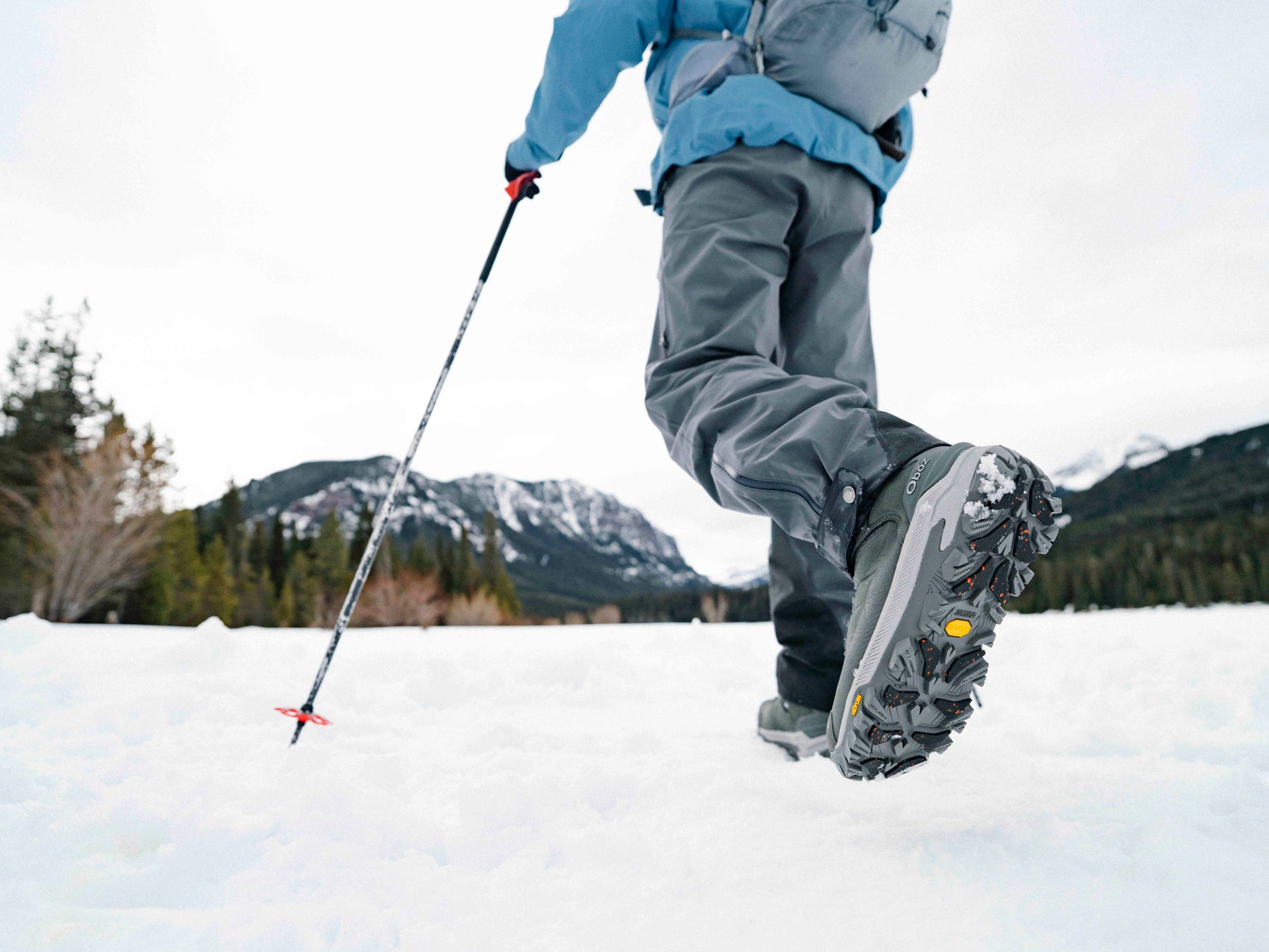 Person hiking in snow wearing Oboz Bangtail winter boots in Charcoal gray with trekking poles, mountainous background.