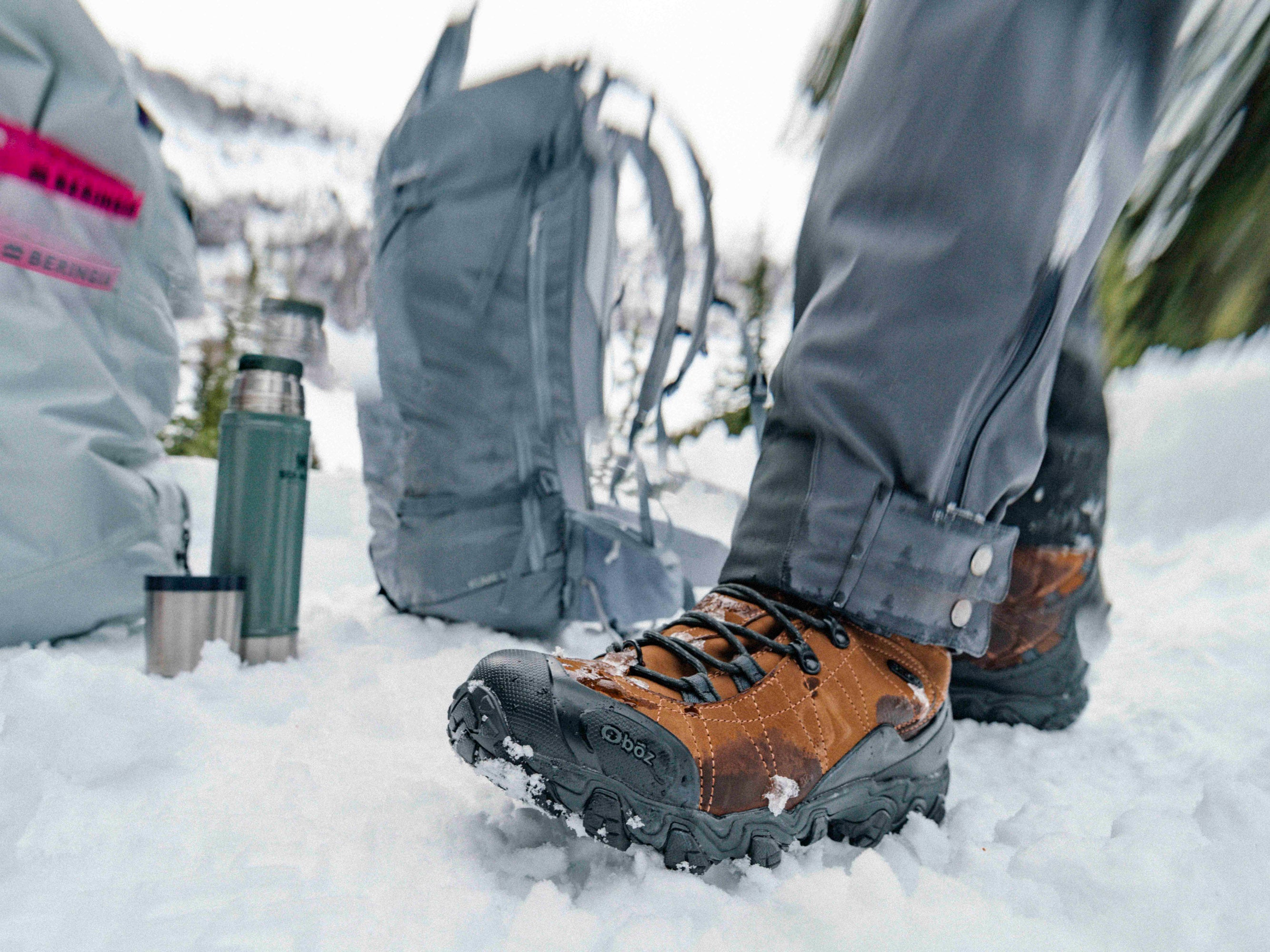 Person wearing brown Oboz Bridger winter hiking boots in the snow with a backpack and thermos in the background.