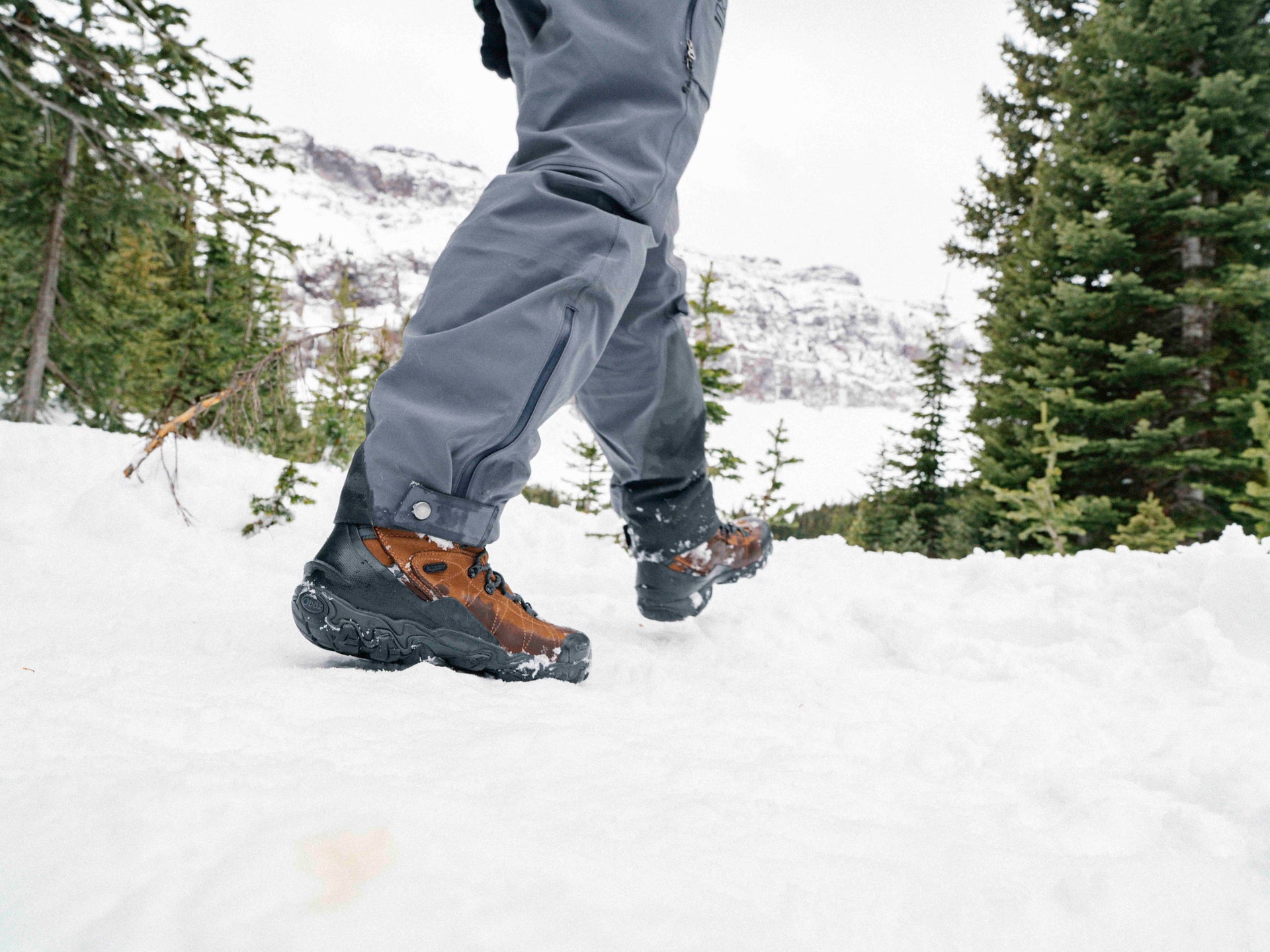 Person wearing brown Oboz Bridger winter boots and gray pants walking on a snowy landscape with trees in the background.