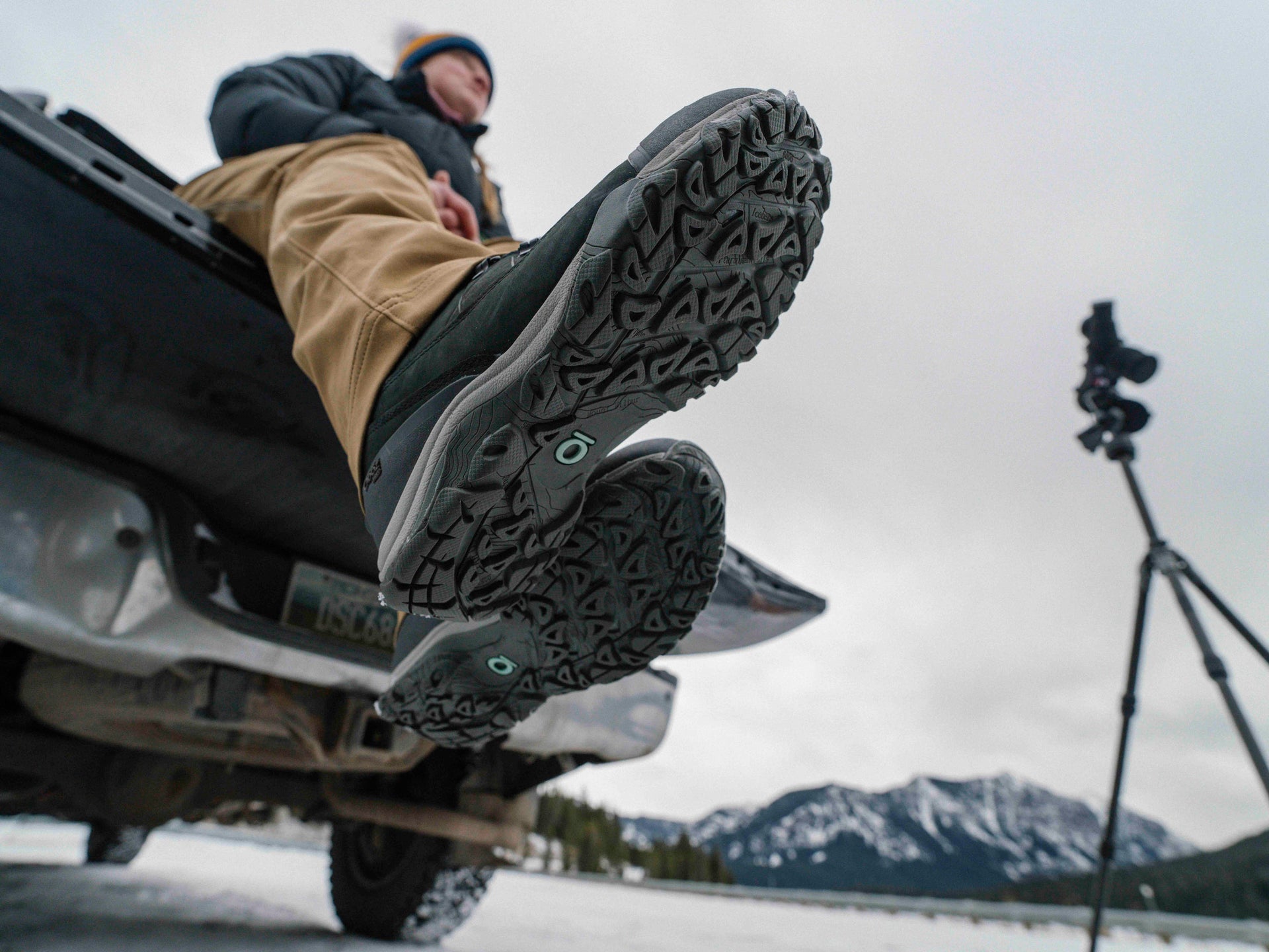 Person sitting on a vehicle's open trunk wearing Oboz winter boots with a mountainous landscape in the background