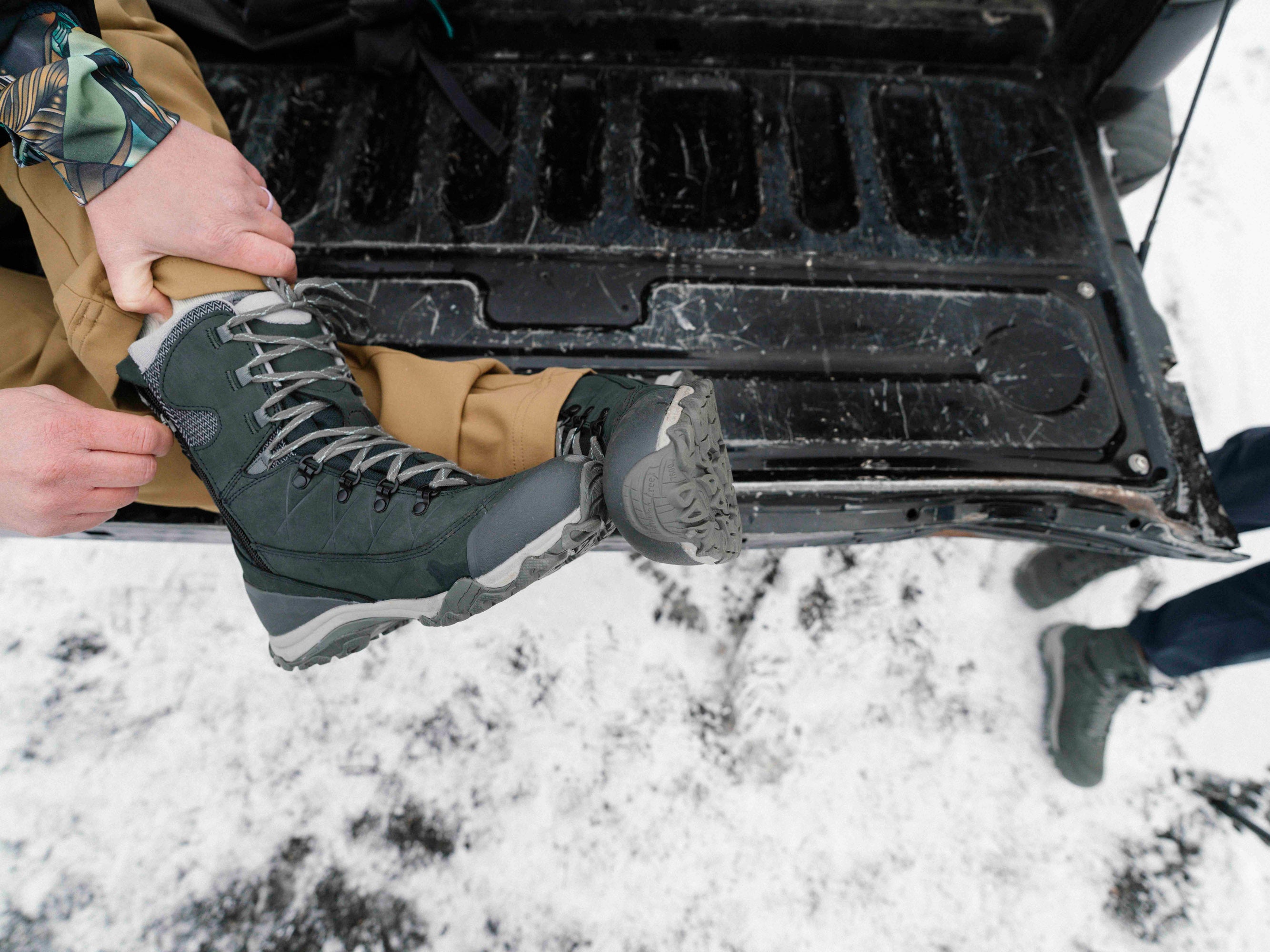 Person putting on black Oboz Ousel Mid Insulated winter boots in front of an open car trunk with snow on the ground.