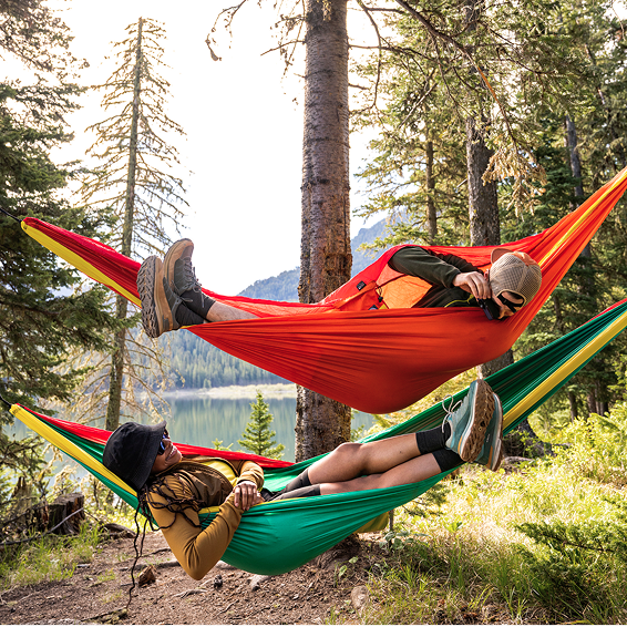 Two people relaxing in colorful hammocks in a forest setting with a lake in the background.
