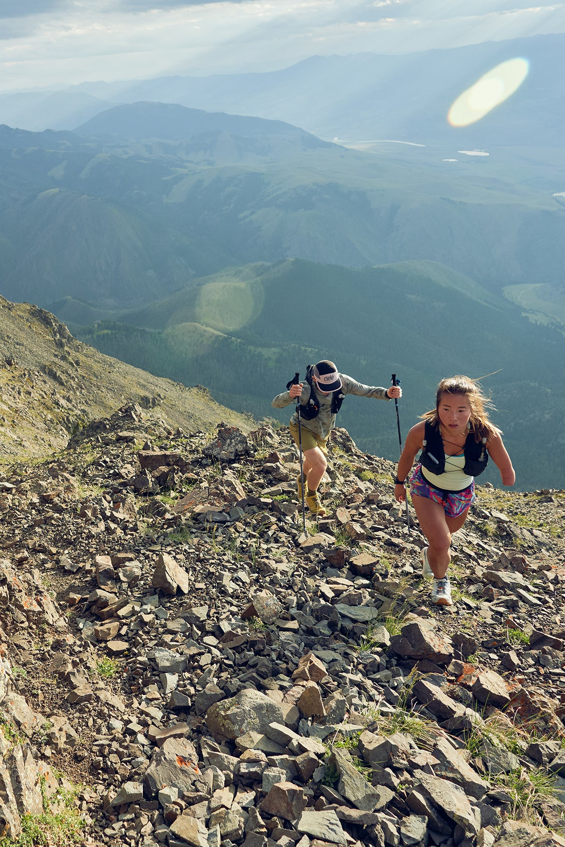 Two hikers on a rocky mountain trail with a scenic view of mountains and valleys.