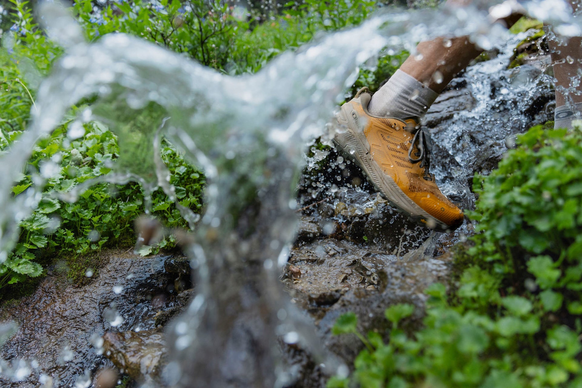 Hiker wearing orange Oboz Katabatic RT waterproof hiking shoes stepping into a stream with water splashing around