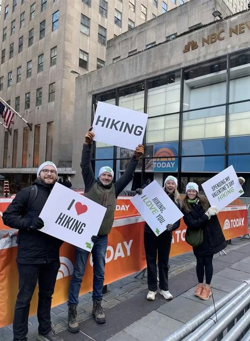People holding hiking-themed signs in front of a building with NBC News branding.