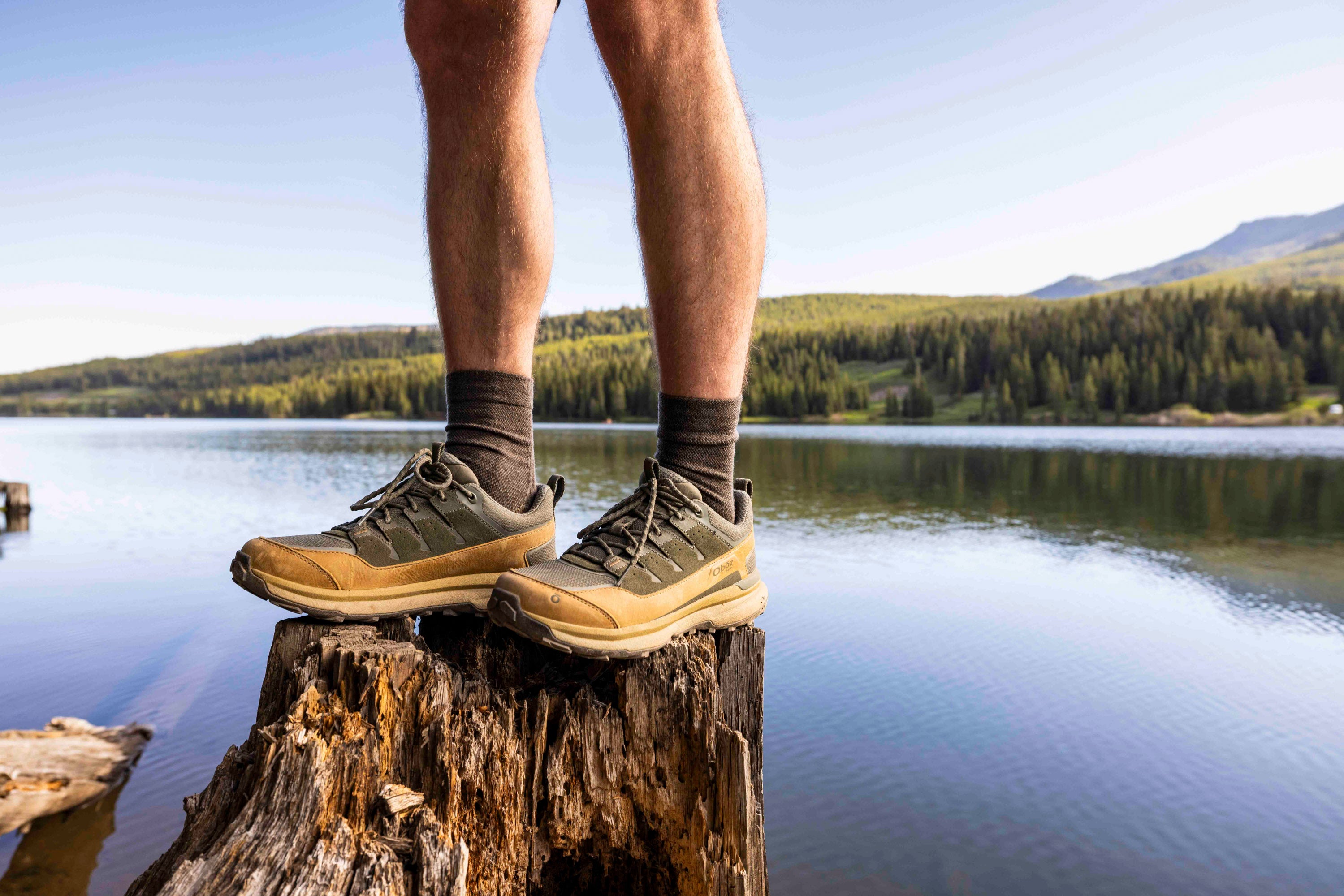 Person wearing Oboz Hyalite hiking shoes standing on a log with a scenic lake and mountains in the background