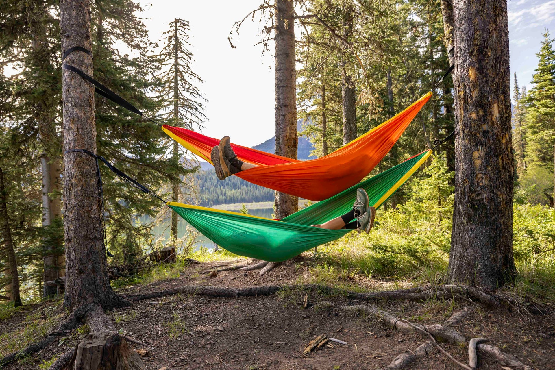 Two people in colorful hammocks in a forest setting with mountains in the background, both wearing Oboz Hyalite hiking shoes