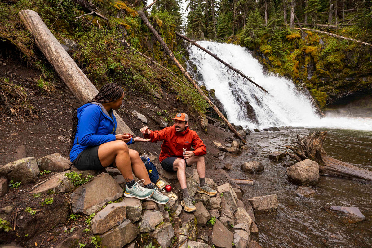 Two people sitting on rocks by a waterfall in a forested area, both wearing Oboz Hyalite hiking shoes