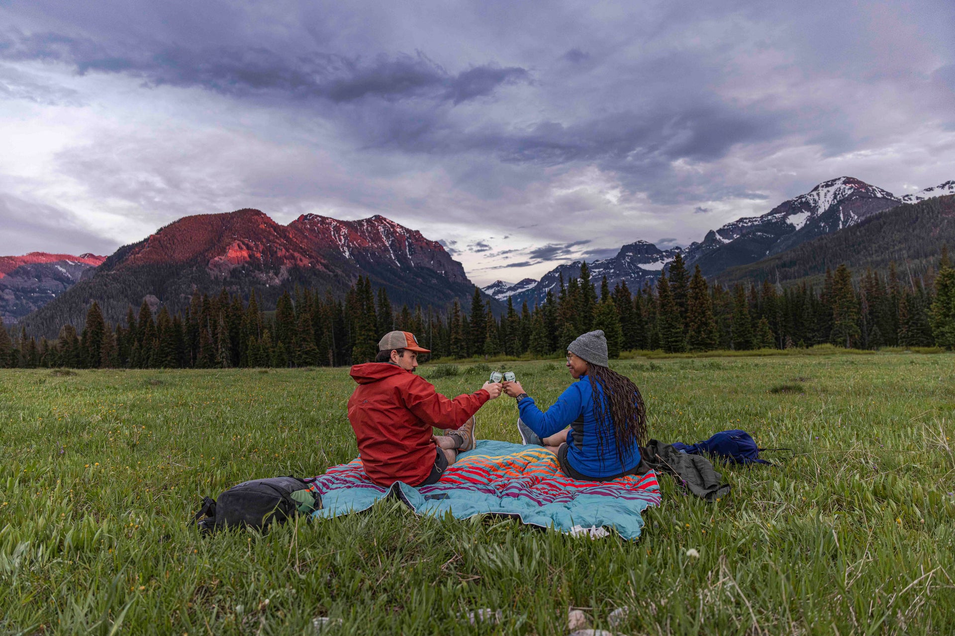 Two people sitting on a colorful blanket in a grassy field with mountains in the background.