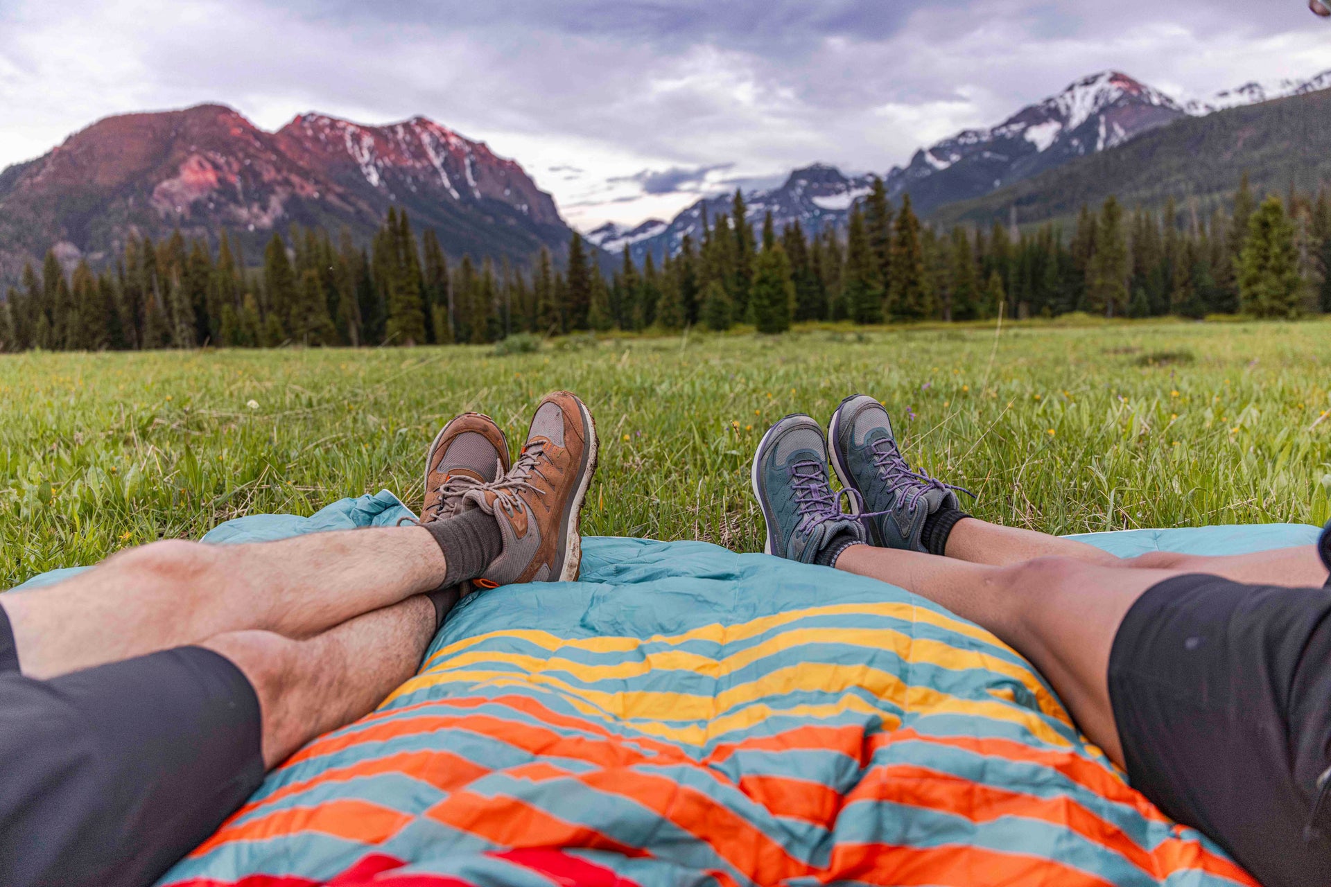 Two people wearing Oboz Hyalite hiking shoes, lying on a colorful blanket in a grassy field with mountains in the background