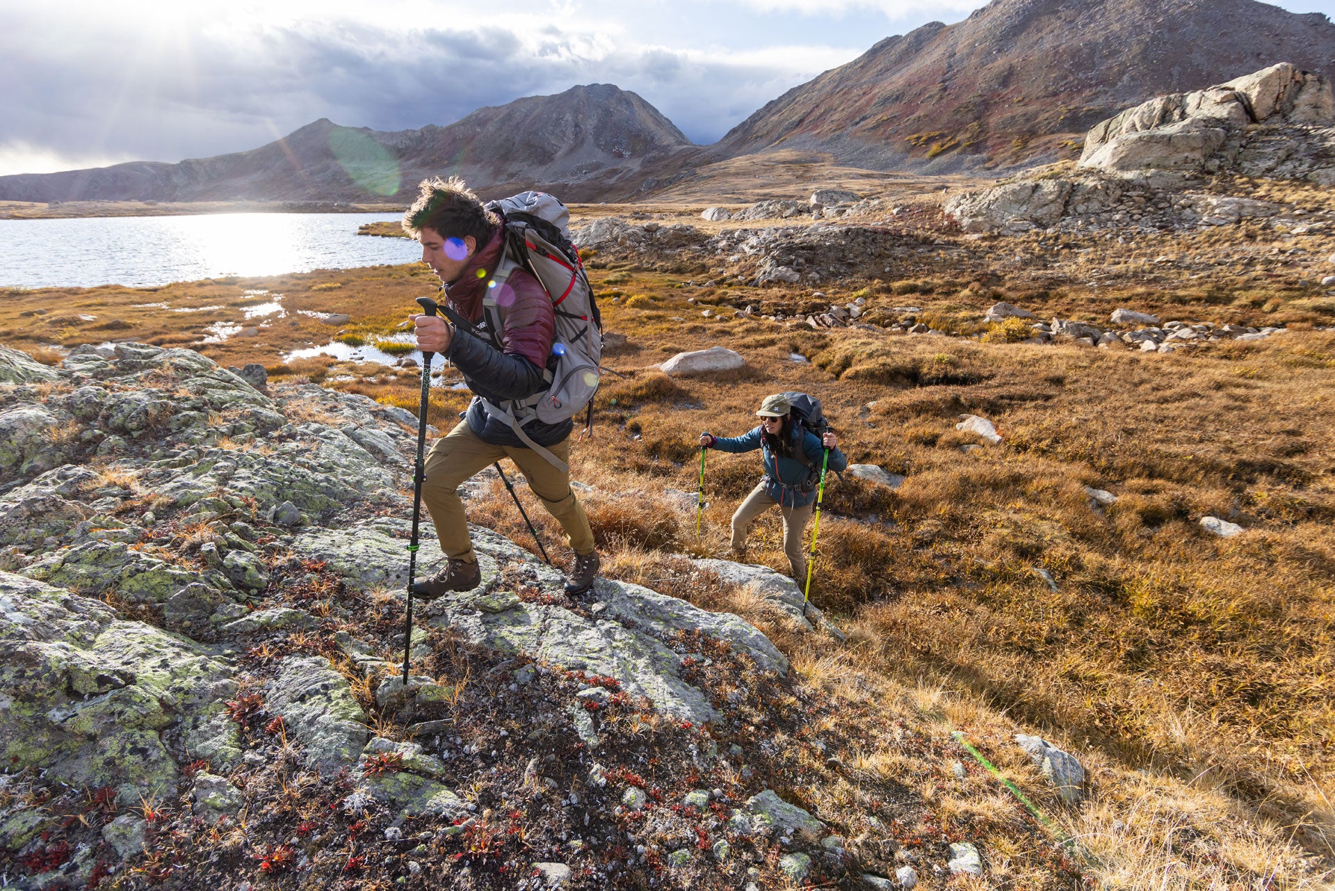 Two hikers with backpacks traversing a rocky landscape with mountains in the background.