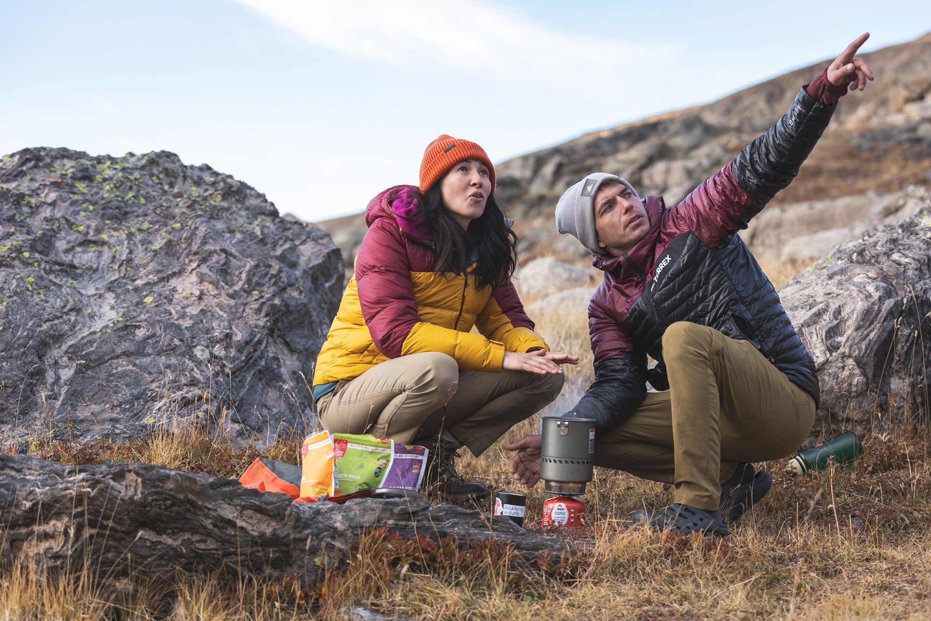 Two people in outdoor gear sitting on rocks with camping equipment in a mountainous area.
