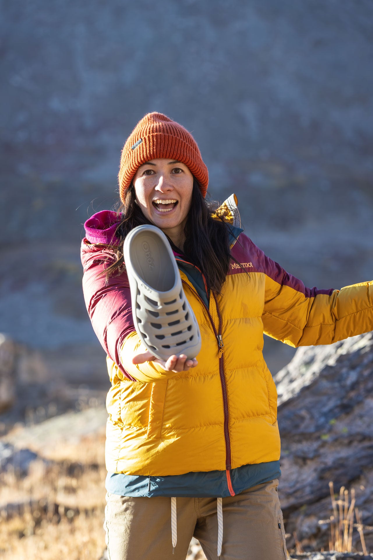 Person wearing a yellow jacket and orange beanie holding up a Oboz Whakata Coast sandal against a mountainous background