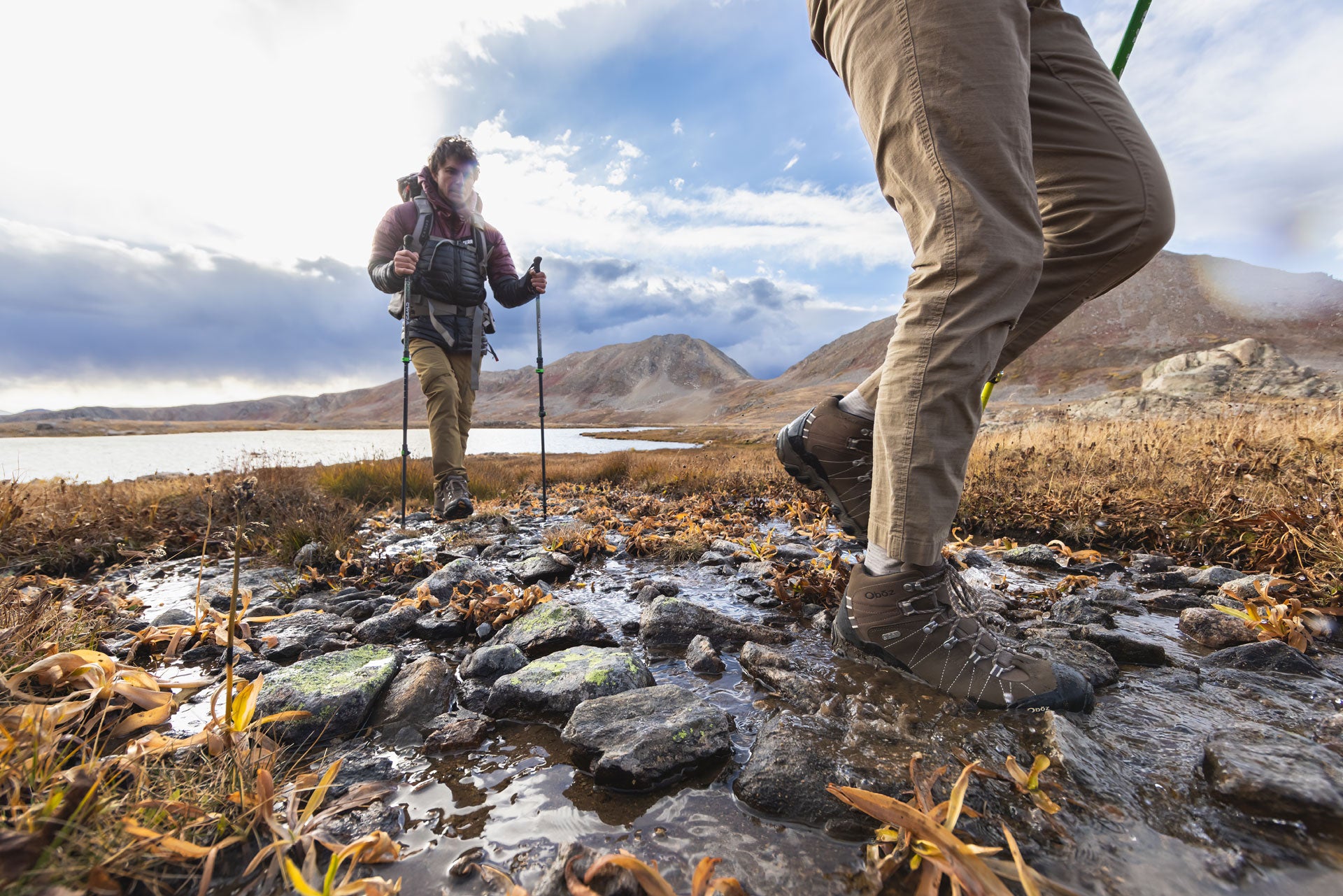 Two hikers wearing Oboz Bridger Mid hiking boots on a rocky trail with mountains and a lake in the background.