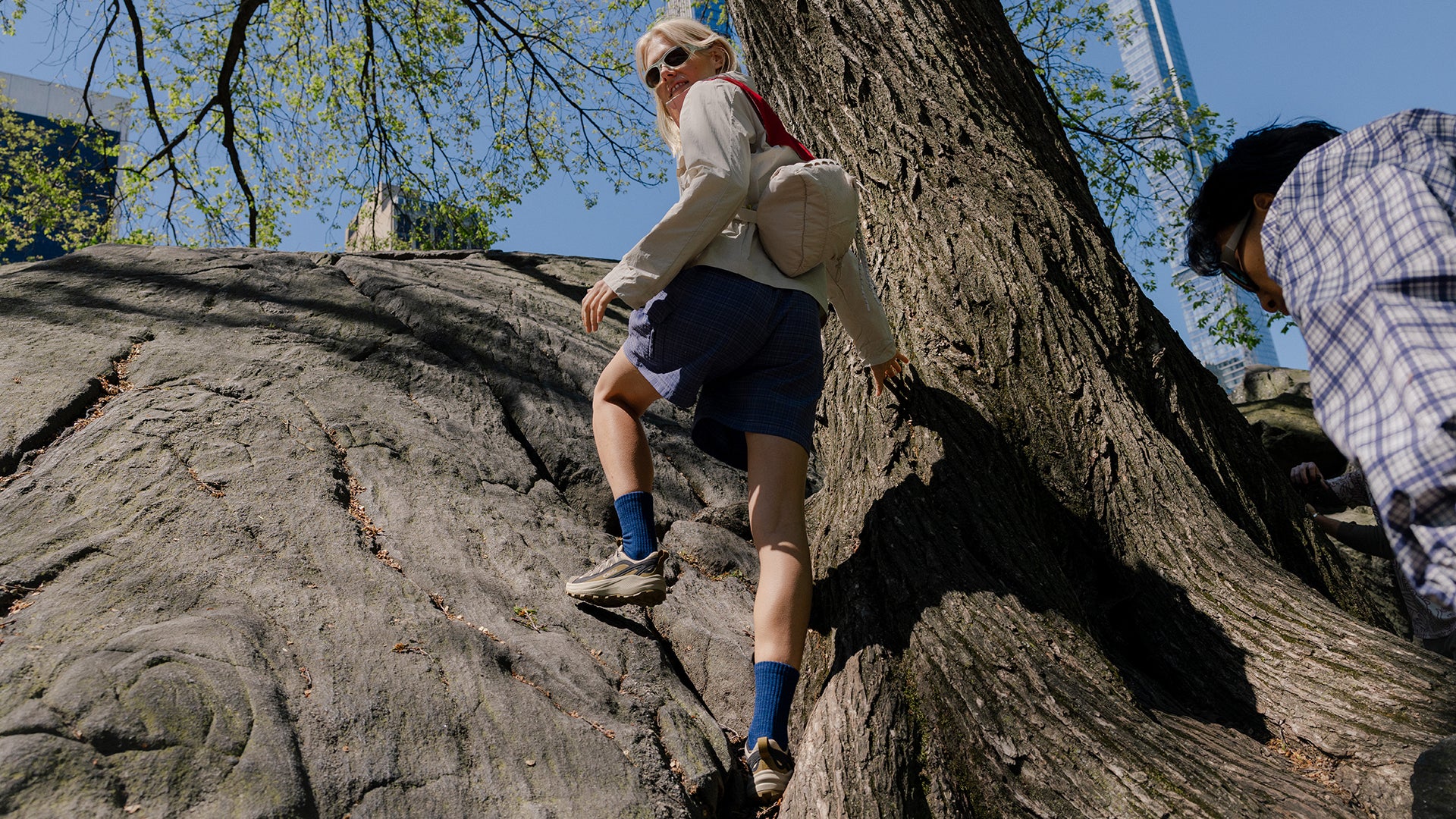 A person wearing a beige long-sleeve shirt, dark blue shorts, blue socks and tan Oboz Crosscut shoes climbs a large tree trunk outdoors under a blue sky.