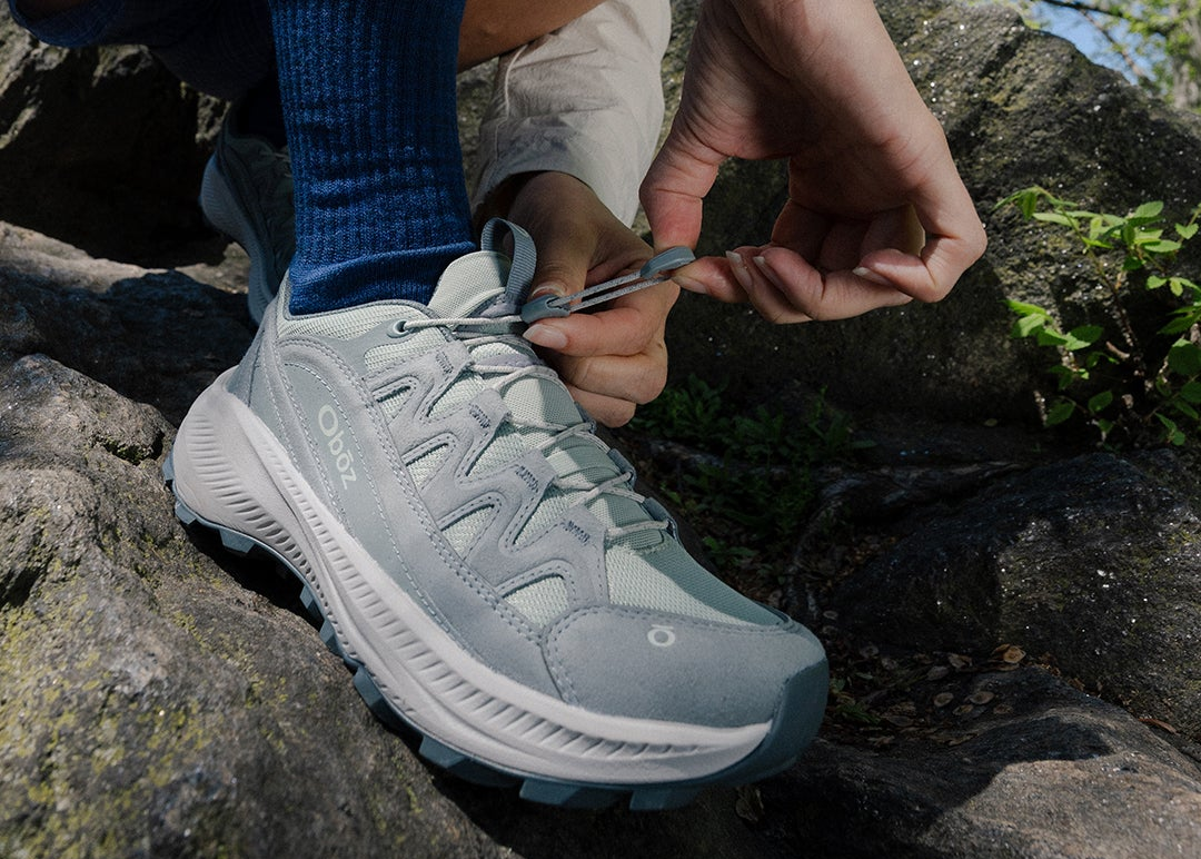 A close-up of a person tying the laces of a light gray Oboz Crosscut hiking shoe while wearing a blue sock, set against a rocky outdoor background.