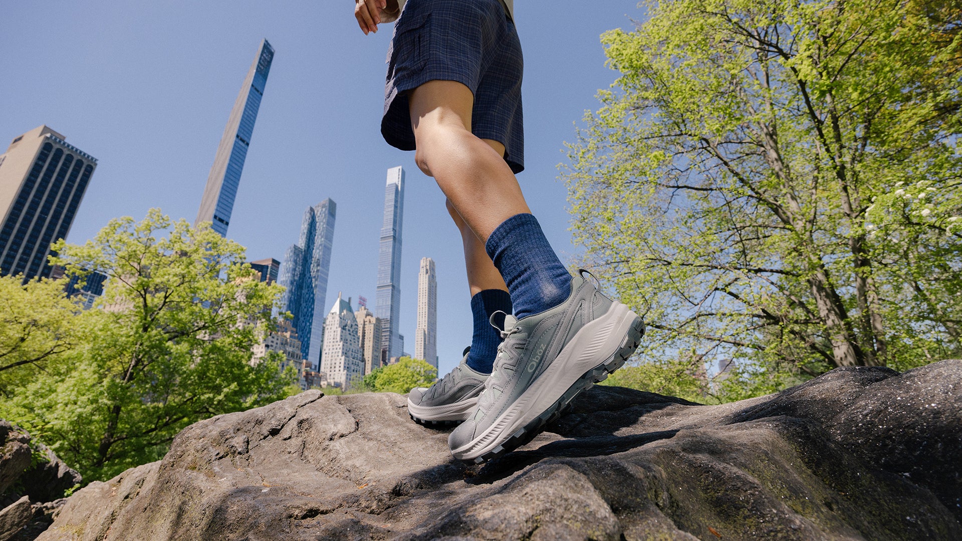 A person's legs and feet wearing gray Oboz Crosscut shoes, blue socks, and dark shorts while climbing a large rock in an outdoor park setting with green trees and a city skyline under a blue sky.