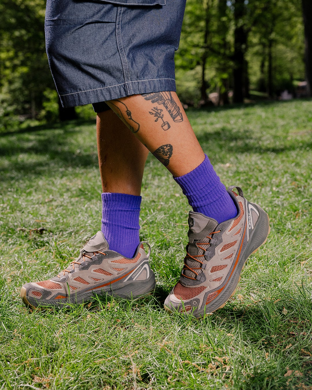 A person standing on grass wearing purple crew socks, Copper colored Oboz Crosscut shoes, and dark denim cargo shorts.