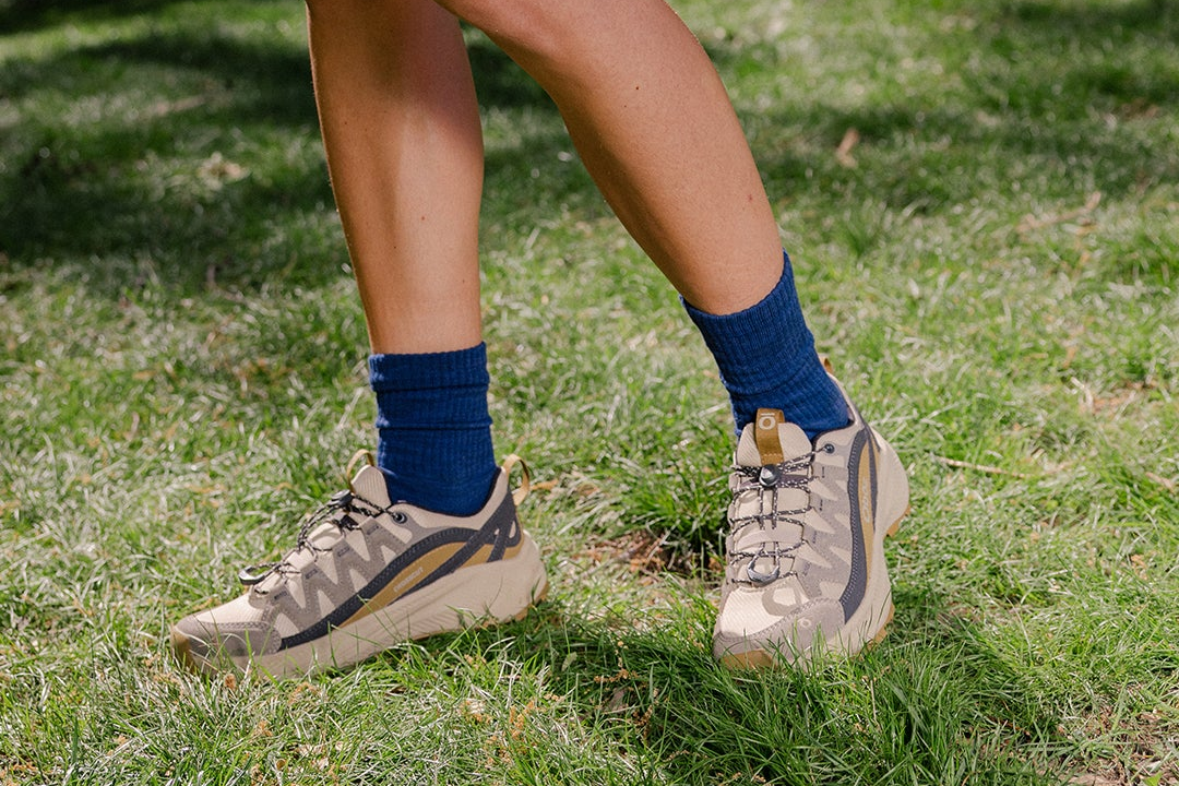 A close-up outdoor shot of a person's legs and feet wearing beige Oboz Crosscut hiking shoes with dark gray accents, paired with dark blue crew socks on a green grassy lawn.