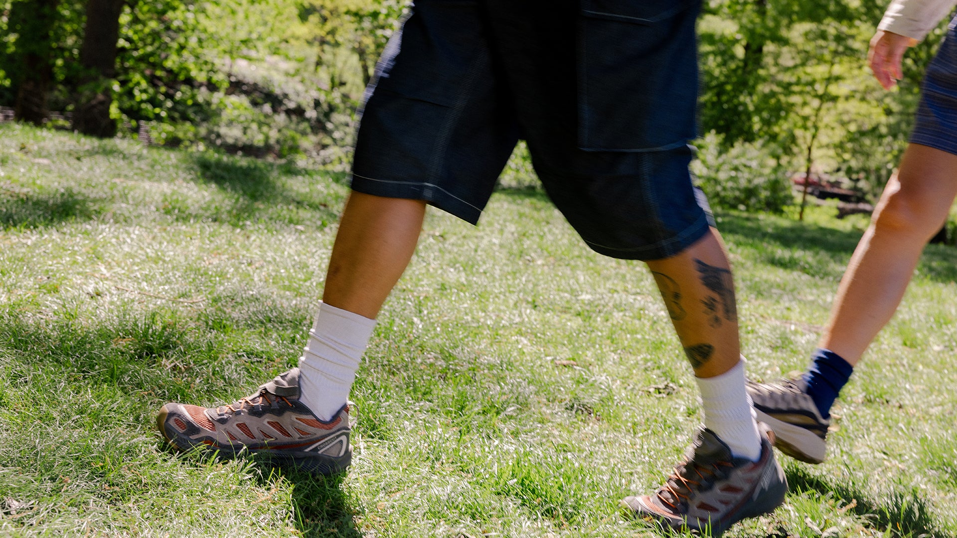 Close-up outdoor shot of a person walking on grass, showing the lower legs wearing dark shorts, white socks, and Oboz Crosscut shoes. 