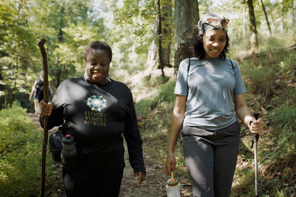 Two women hiking in a forest, one wearing a black shirt with 'Refugee Women's Network' text.