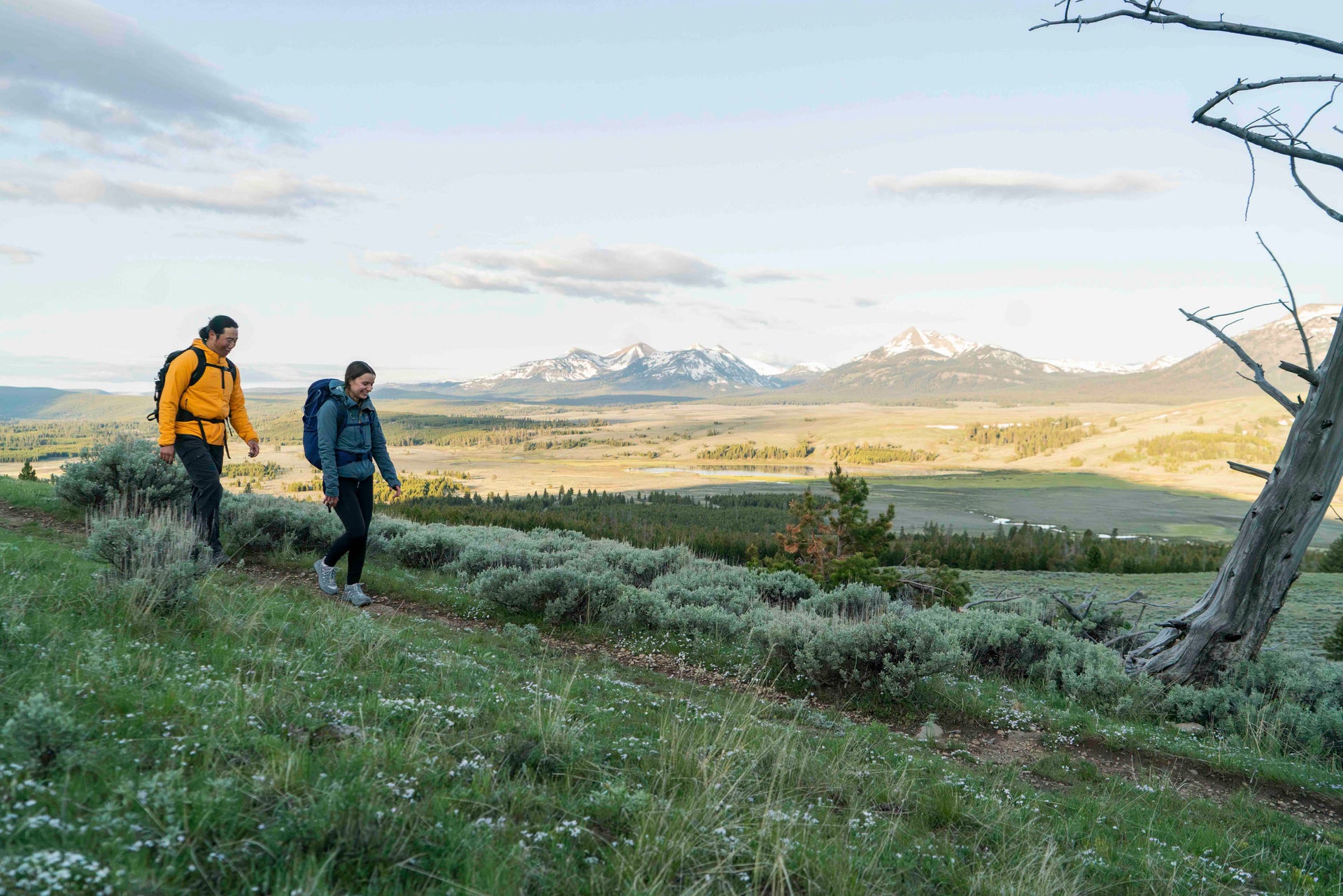Two hikers with backpacks walking on a trail with mountains in the background