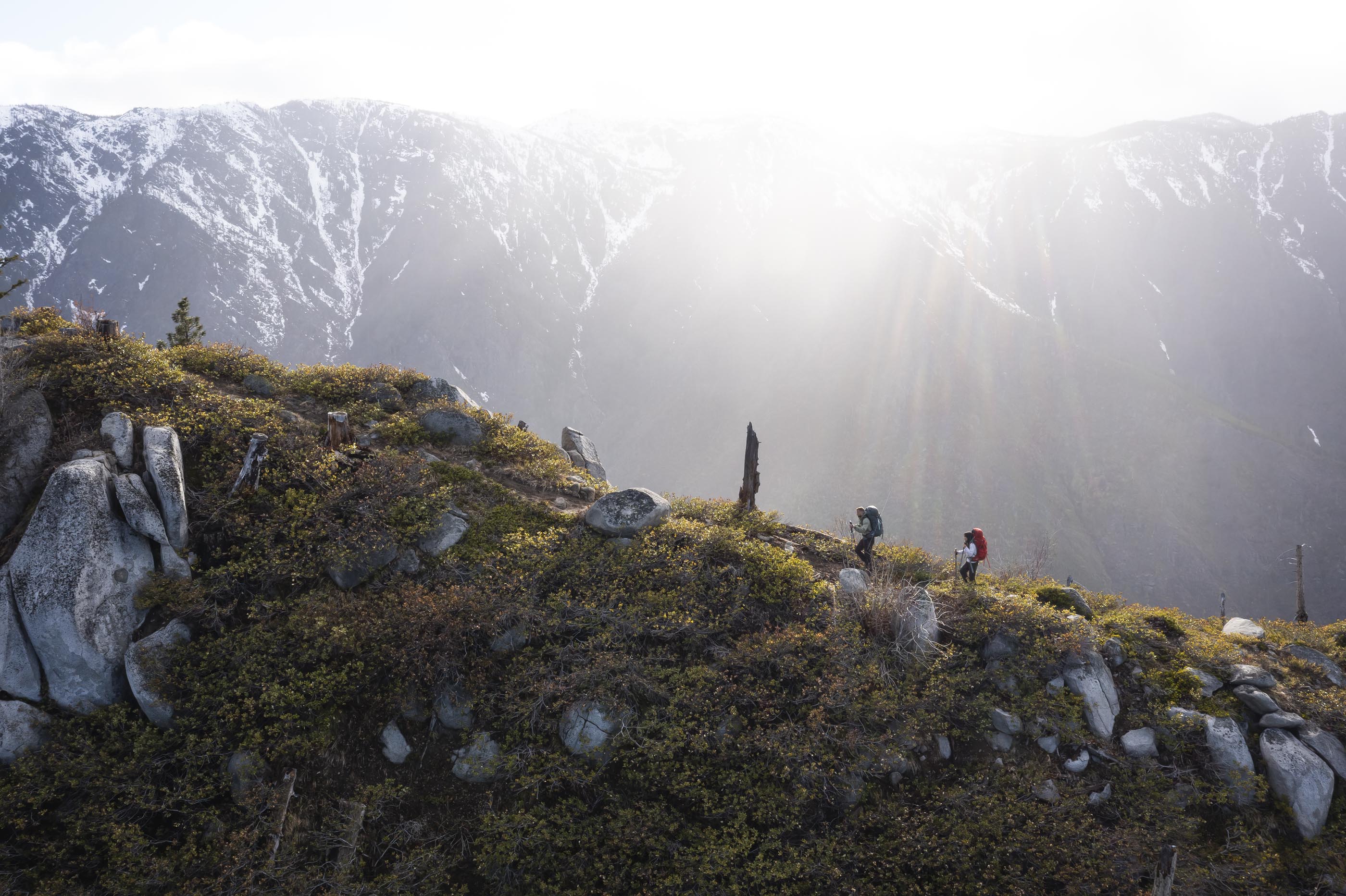 Two people hiking on a mountain trail with snow-capped peaks in the background.
