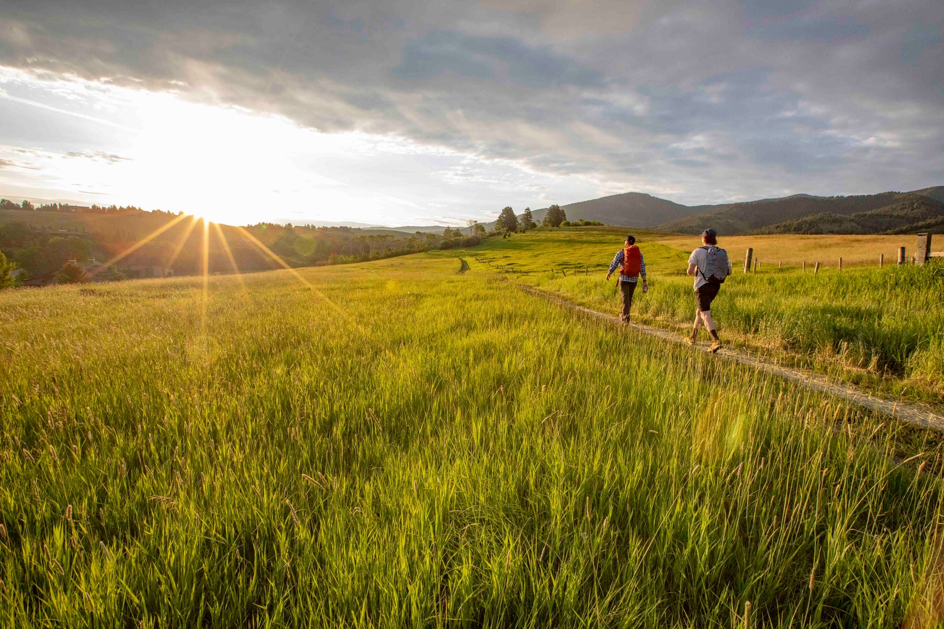 Two people walking along a dirt path in a grassy field with mountains in the background.