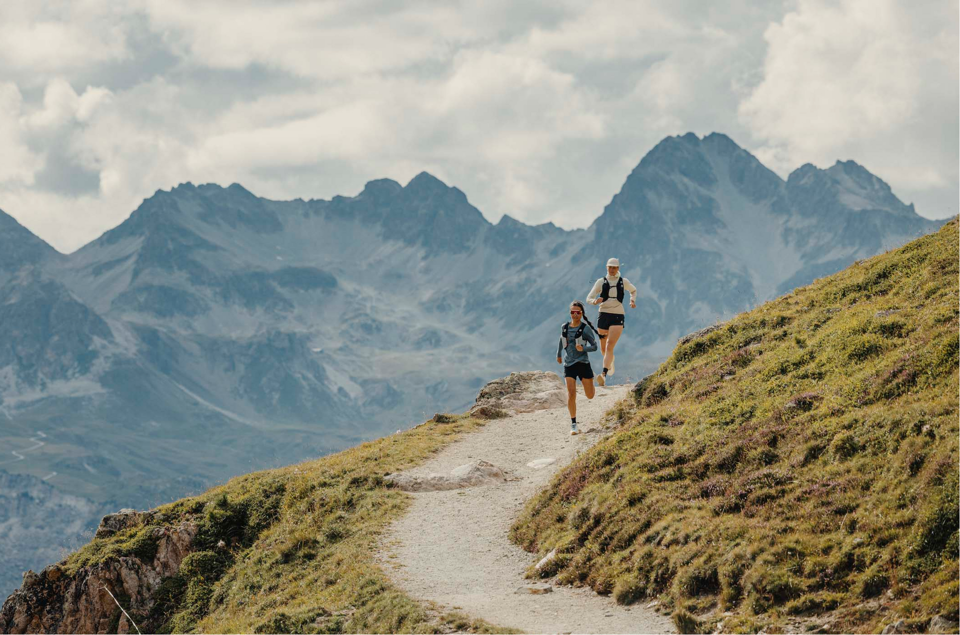Two people running on a mountain trail with a scenic mountain landscape in the background.