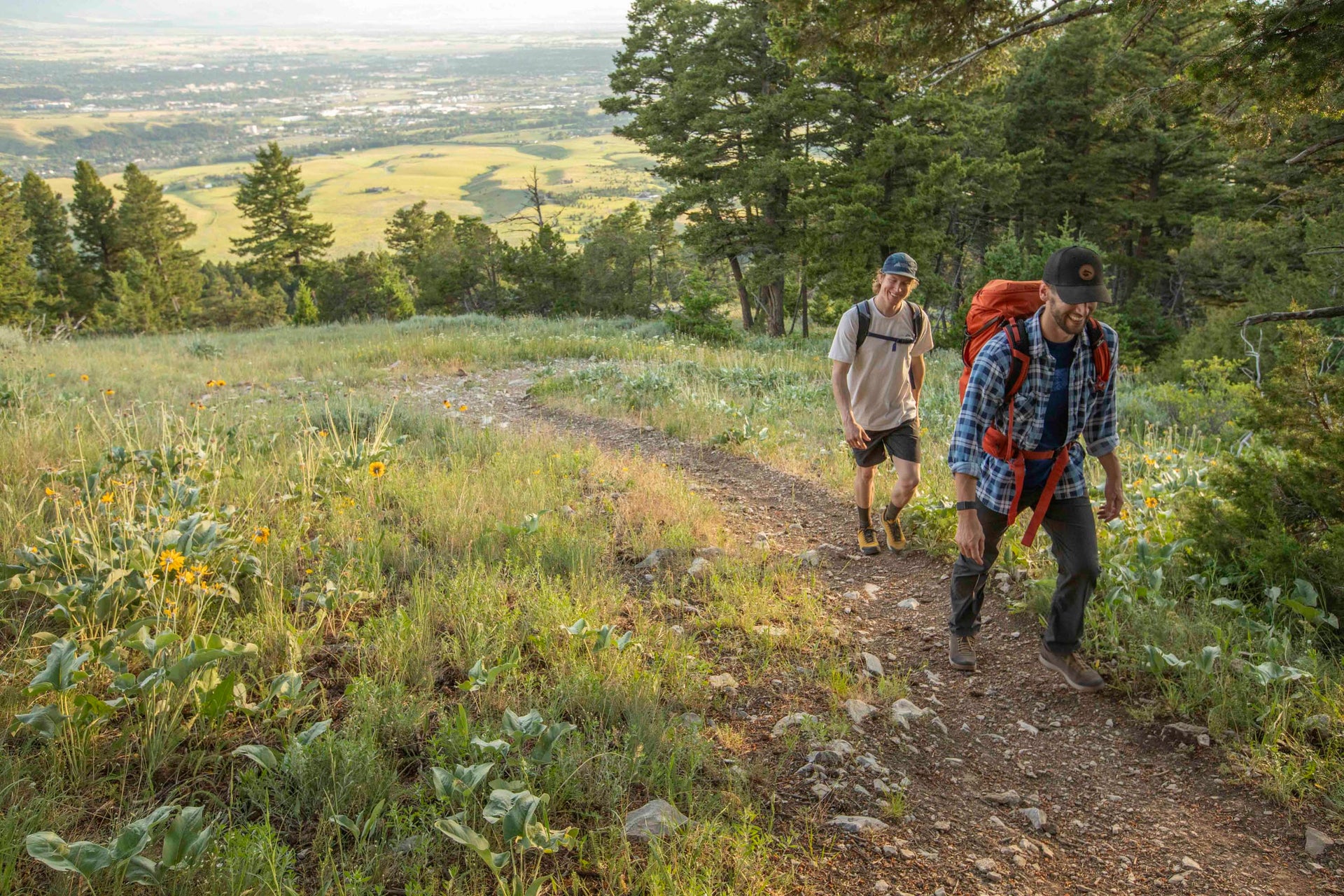 Two hikers wearing Oboz Bozeman II hiking shoes, on a trail with a scenic view of trees and fields.