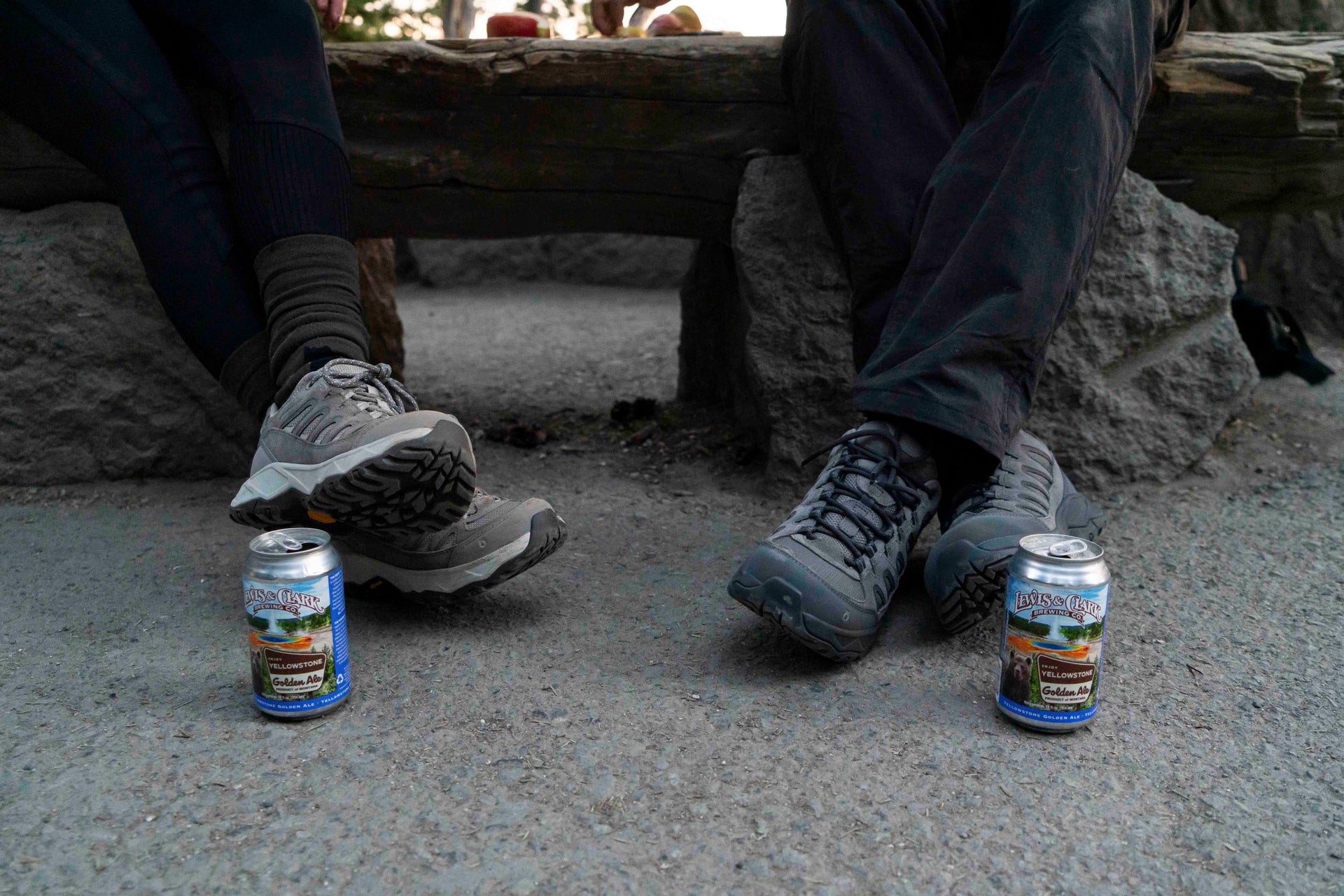 Two cans of beer on the ground with people sitting on a stone bench in the background wearing Oboz Sawtooth Ascent hiking boots