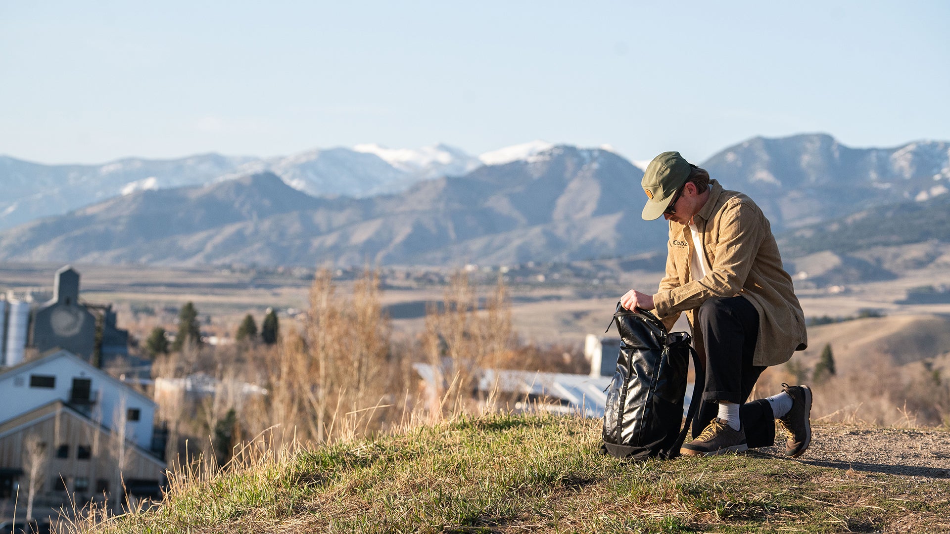 A man in a tan jacket and green cap kneels on a grassy hillside, packing gear into a black backpack, with a scenic mountain valley in the background.