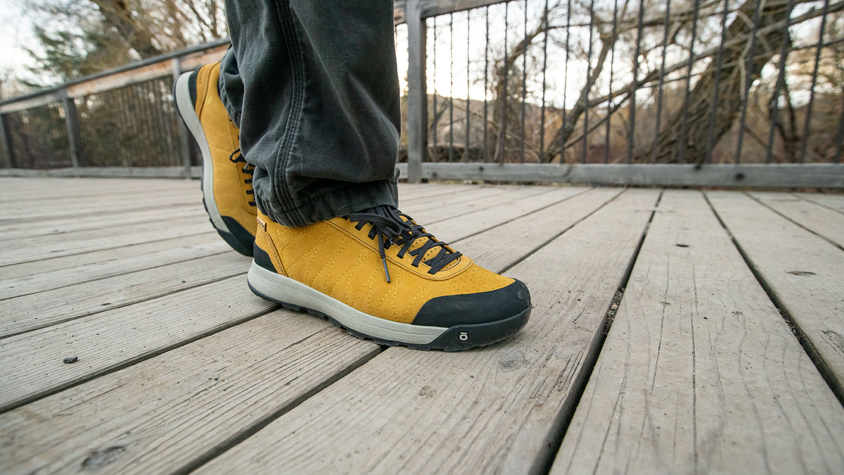 A person wearing yellow Oboz Bozeman II shoes with dark gray pants, standing on a wooden bridge outdoors.